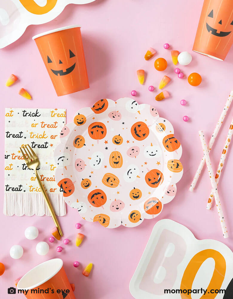A pink Halloween party table featuring Momo Party's hey pumpkin tableware including a round pink scallop edged plates with cute jack o lantern, an orange jack o lantern party cups, pink trick or treat fringe napkins, boo shaped plates and pink pumpkin reusable straws. With pink Halloween treats like orange gum balls, candy cones, pink candies, it makes a great table setting inspiration for a cute girly kid's pink Halloween party table setting.