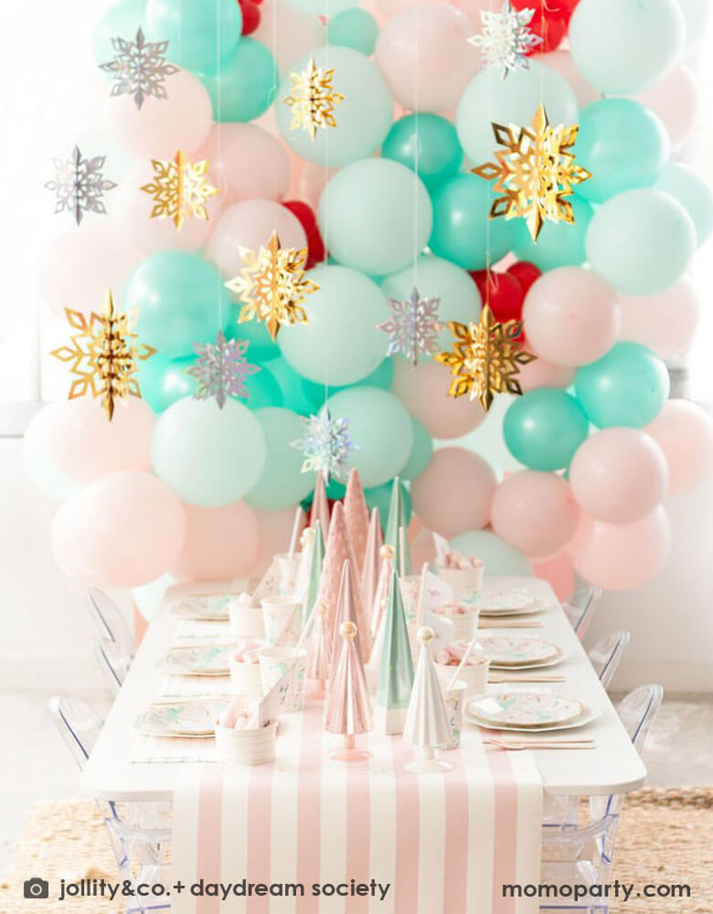 A pink Christmas party table featuring Momo Party's magical unicorn plates and cups by Daydream Society. A few ceramic Christmas tree decorations in mint and pink are used as the centerpiece on a pink white striped table runner. In the back of the table there's a balloon wall with pastel pink and mint balloons adorned with gold and silver snowflake hanging decorations, all makes it a whimsical kid's Christmas party inspo. 