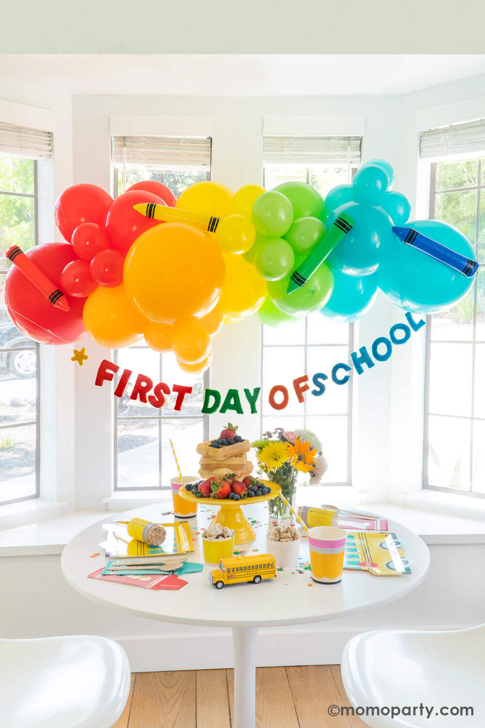 A bright, modern kitchen bay window in the morning decorated with Momo Party's first day of school balloon garlands in fun and bright colors. Underneath is a "first day of school" felt banner by Studio Pep. Along with a table filled with delicious breakfast food including stacks of Belgium waffles, cereals, croissants and muffins with school themed tableware from Momo Party. This makes a great inspiration for a back to school celebration for kids at home.