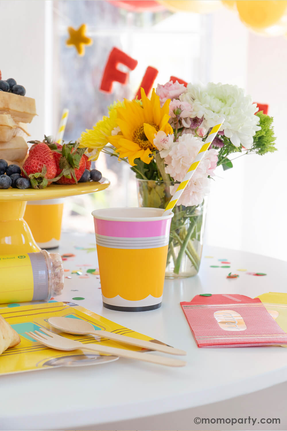 A first day of school breakfast celebration party table by Momo Party featuring pencil shaped party cups, school bus shaped plates, and colorful crayon shaped napkins. Along with the felt "first day of school" party banner in the back and fresh floral arrangement as the centerpiece, it makes great inspiration for an at home back to school party.