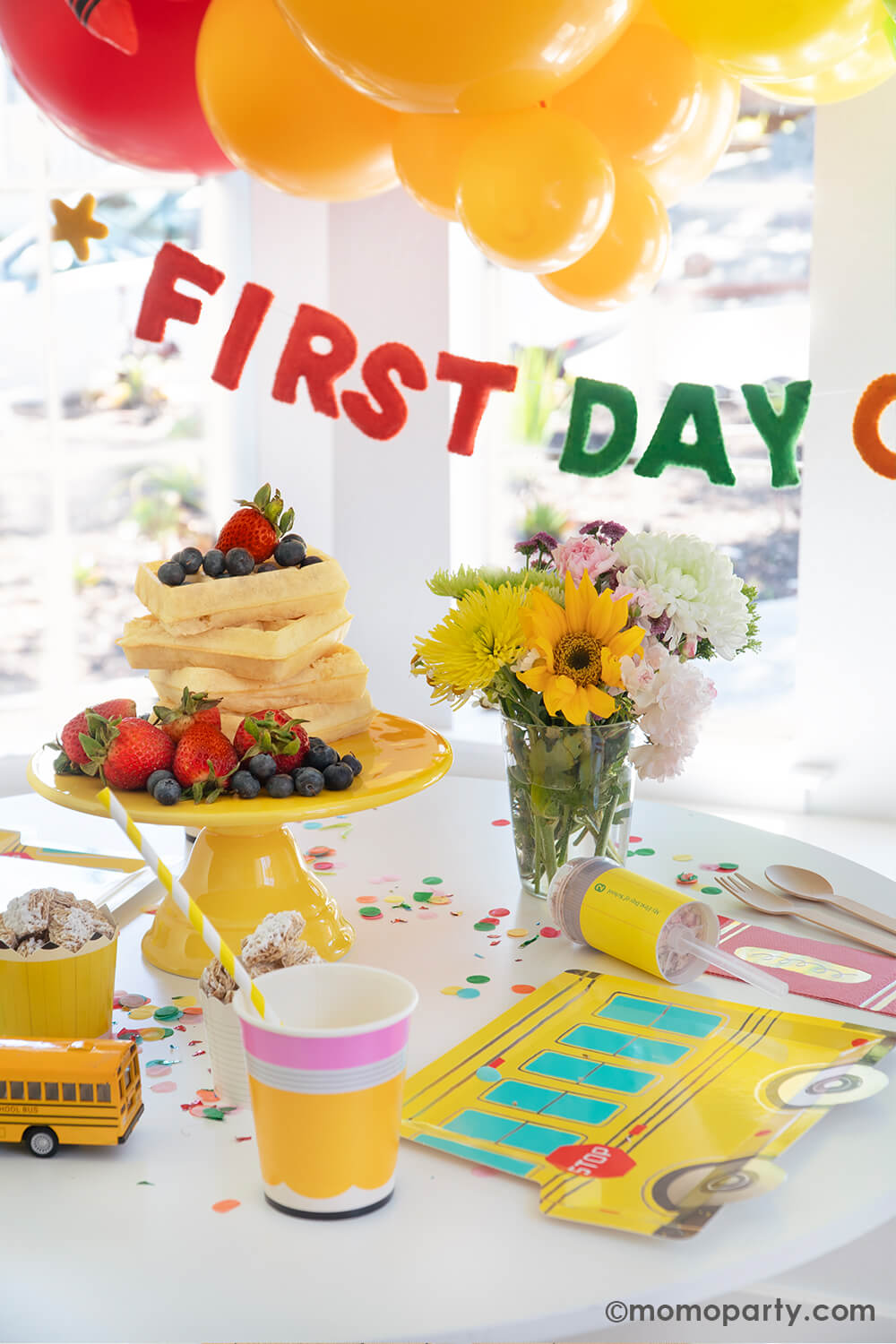 A bright, modern kitchen bay window in the morning decorated with Momo Party's first day of school balloon garlands in fun and bright colors. Underneath is a "first day of school" felt banner by Studio Pep. Along with a table filled with delicious breakfast food including stacks of Belgium waffles, cereals, croissants and muffins with school themed tableware from Momo Party. This makes a great inspiration for a back to school celebration for kids at home.