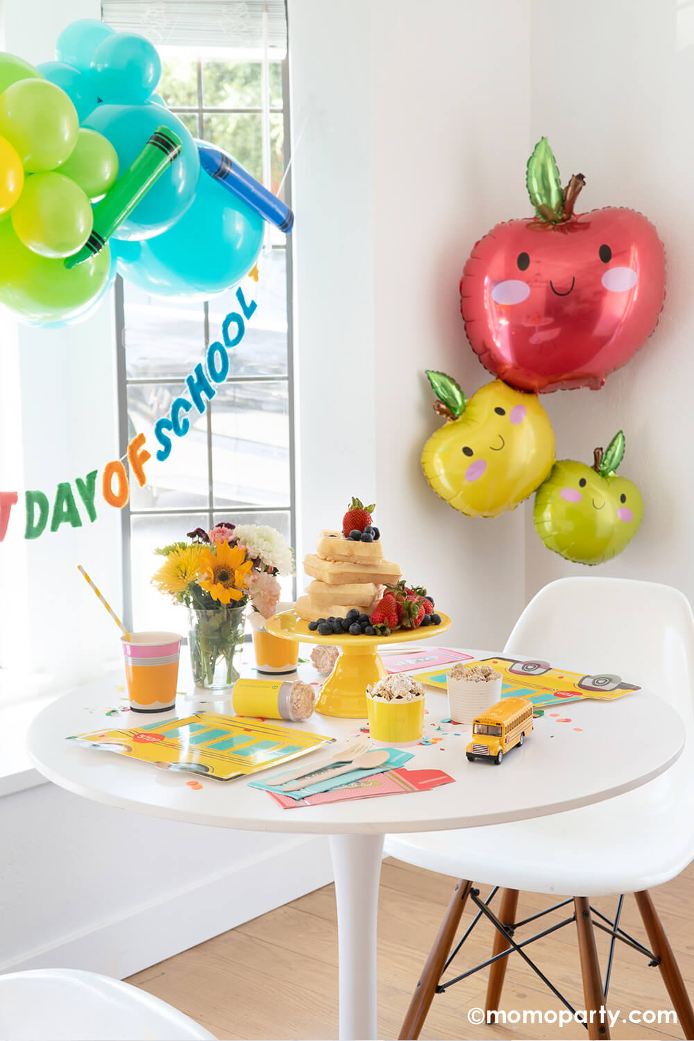 A kitchen table area decorated with back to school themed balloons in fun, bright colors and an apple stacker foil balloon in a adorable illustration appealing to kids. On the table there is a stack of Belgium waffles as the "breakfast cake" and multiple school themed tableware from momoparty.com. All in all creates a great look and inspiration for kid's back to school celebration or party.