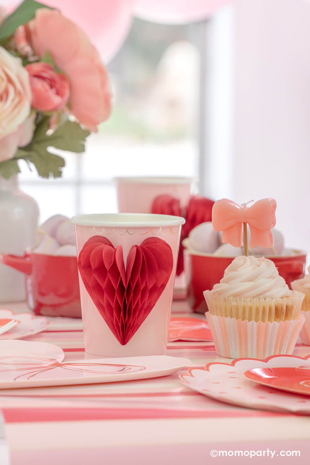 A close-up detail of Momo Party's sweet Pink and Red Bow-Themed Valentine's Day Party table. Highlighting Meri Meri Honeycomb Heart Cups, these cups feature charming 3D honeycomb red hearts on pink paper, adorned with gold foil details. Accompanied by a Pink Bow Candle atop a cupcake and sweets in the red Mini Cocotte, with a flower vase behind them. All elegantly arranged on a red-striped paper runner. Such a cute idea for elevating your Valentine's celebration or Galentine's Day with your besties.