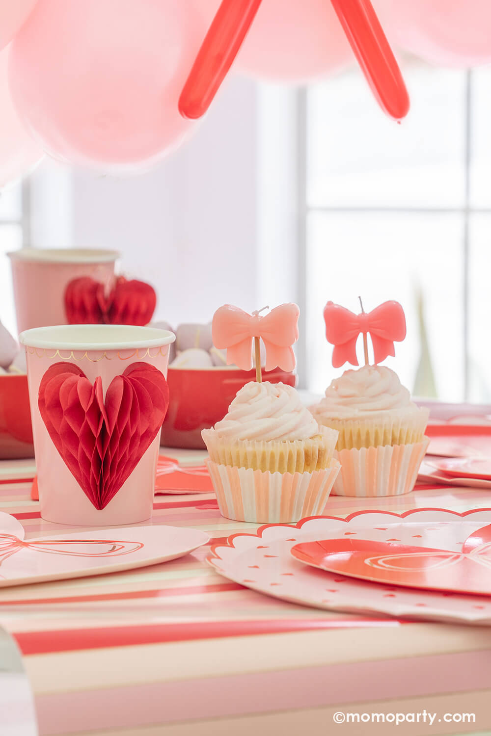 A close-up detail of Momo Party's sweet Pink and Red Bow-Themed Valentine's Day Party table. Highlighting Meri Meri Honeycomb Heart Cups, these cups feature charming 3D honeycomb red hearts on pink paper, adorned with gold foil details. Accompanied by a Pink Bow Candles atop a cupcakes and sweets in the red Mini Cocotte, with a flower vase behind them. All elegantly arranged on a red-striped paper runner. Such a cute idea for elevating your Valentine's celebration or Galentine's Day with your besties.