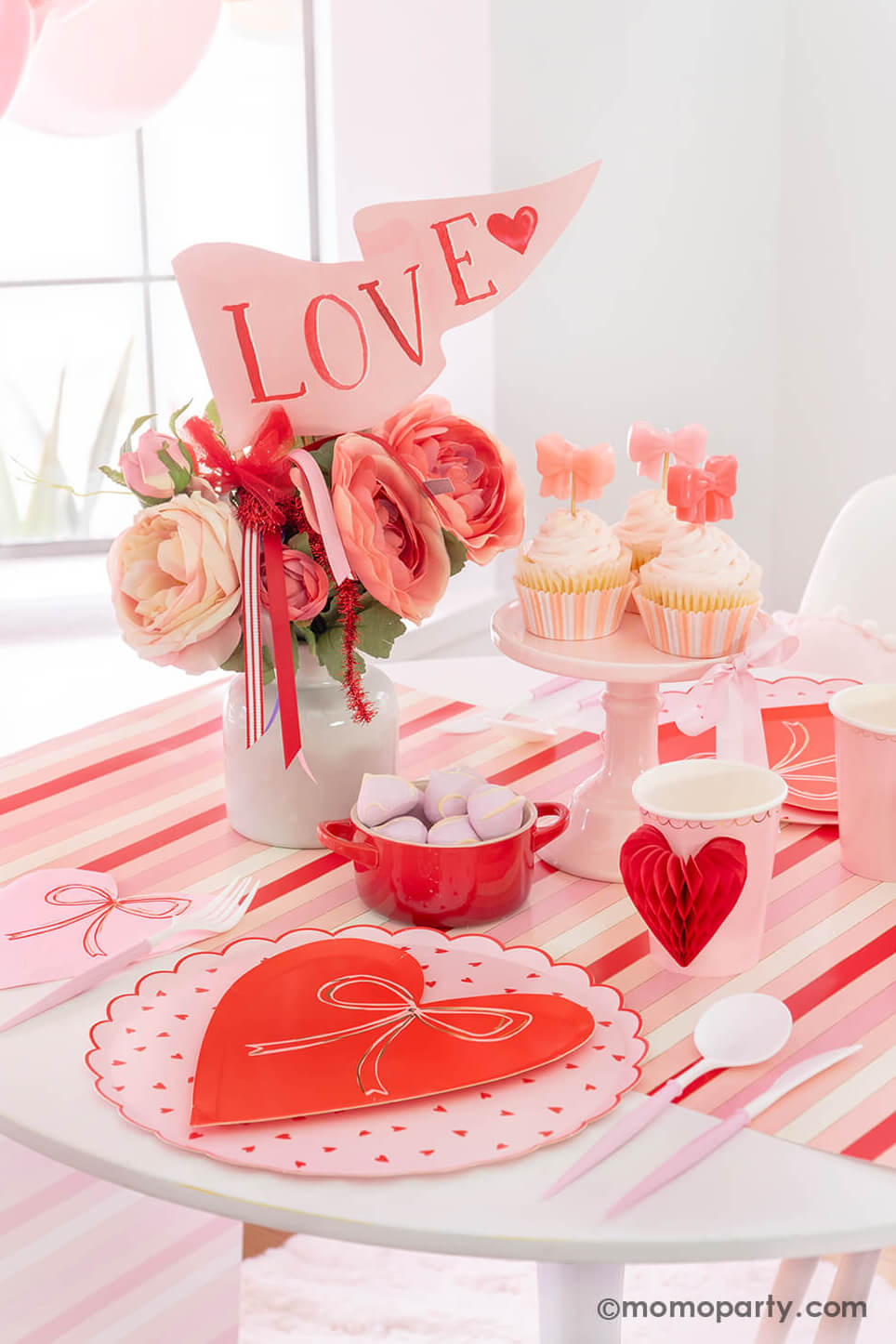 Momo Party presents A Bow-Themed Valentine's Day Table: This Bow-tastic Table Setting is filled with Meri Meri Heart Pattern Dinner Plates layered with Bow Plates and matching napkins, Honeycomb Heart Cups, Blush and White Cutlery Set, Pink Bow Candles on cupcakes, and a Love Party Pennant in the flower vase, all elegantly arranged on a red-striped paper runner. What a modern and cute idea for your Valentine's or Galentine's celebration with besties."