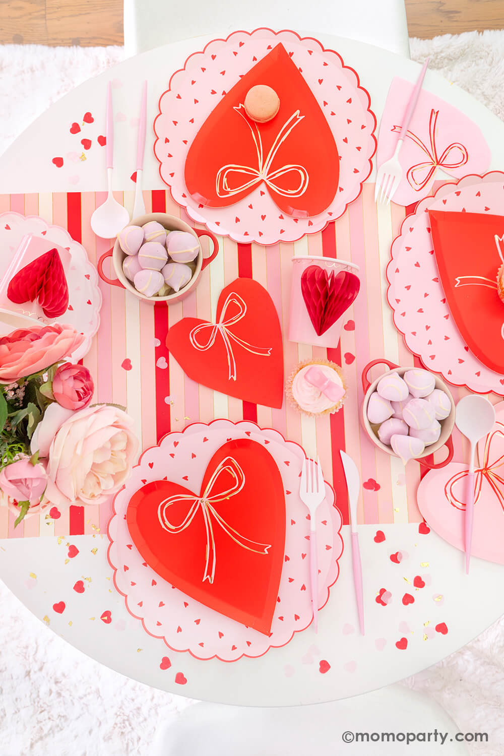Top view of Momo Party's sweet Pink and Red Bow-Themed Valentine's Day Party table. Featuring Meri Meri Heart With Bow Plates layered with Heart Pattern Dinner Plates, Heart With Bow Napkins, Honeycomb Heart Cups, Blush and White Cutlery Set, Pink Bow Candles atop cupcakes, and sweets in the red Mini Cocotte. All elegantly arranged on a red-striped paper runner, sprinkled around Kisses Artisan Confetti. Such a cute idea for elevating your Valentine's celebration or Galentine's Day with your besties
