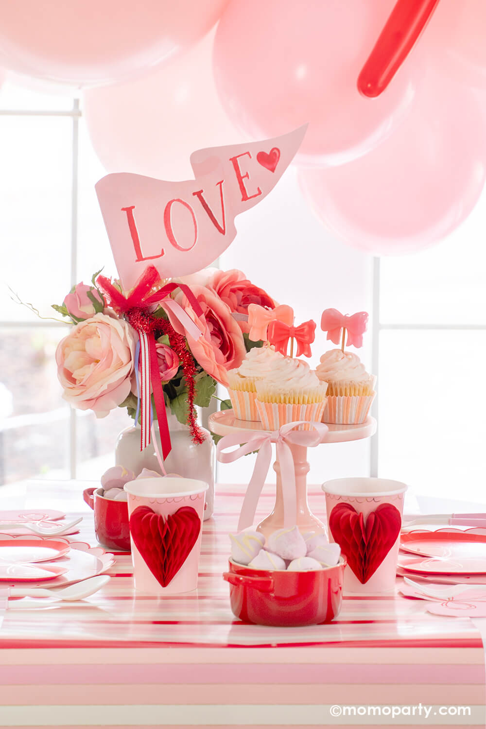 Momo Party presents A Bow-Themed Valentine's Day Party Set up. This Bow-tastic Table Setting is filled with heartwarming details, featuring Meri Meri Heart Pattern Dinner Plates layered with Bow Plates and matching napkins, Honeycomb Heart Cups, Pink Bow Candles atop cupcakes, and a Love Party Pennant adorning the flower vase, all elegantly arranged on the red-striped paper runner. It's a modern and cute idea to elevate your Valentine's celebration or Galentine's Day with your besties. 
