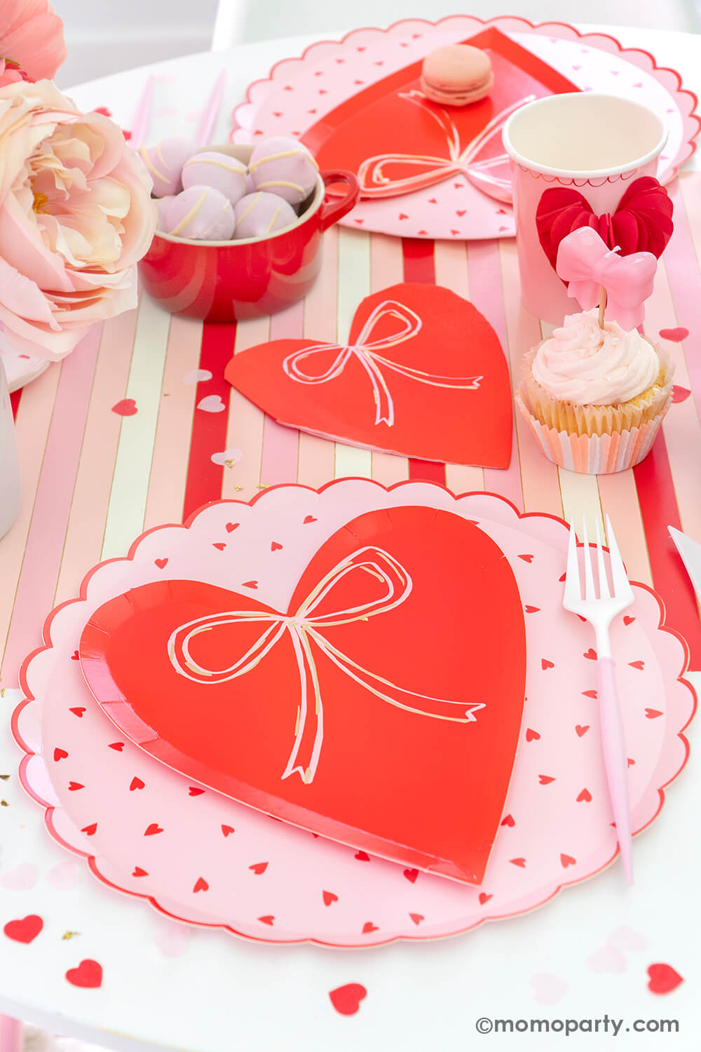 Momo Party presents a sweet Pink and Red Bow-Themed Valentine's Day Party table. Featuring Meri Meri Heart With Bow Plates layered with Heart Pattern Dinner Plates, Heart With Bow Napkins, Honeycomb Heart Cups, Blush and White Cutlery Set, Pink Bow Candles atop cupcakes, and sweets in the red Mini Cocotte. All elegantly arranged on a red-striped paper runner, sprinkled around Kisses Artisan Confetti. Such a cute idea for elevating your Valentine's celebration or Galentine's Day with your besties.
