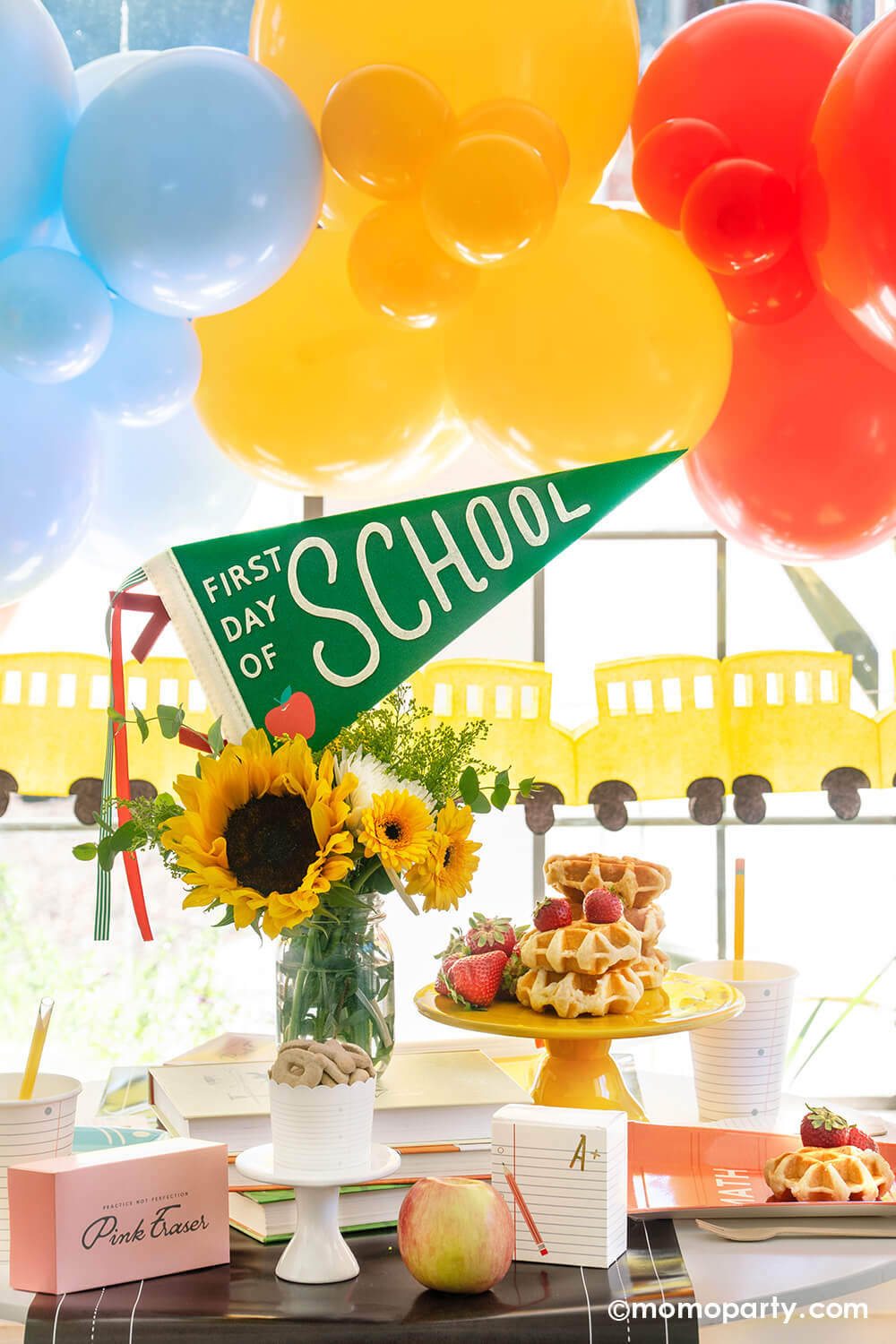 An adorable first day of school breakfast table setting featuring school themed party goods from Momo Party including the first day of school felt party pennant, chalkboard table runner, pink eraser shaped treat boxes, notebook shaped treat boxes, text book shaped plates, notebook paper cups and pencil straws. There are also some waffles stacked and strawberries on a cake stand. Behind the table is a festive balloon garland and school bus party banner, a perfect set up for your child's first day of school.