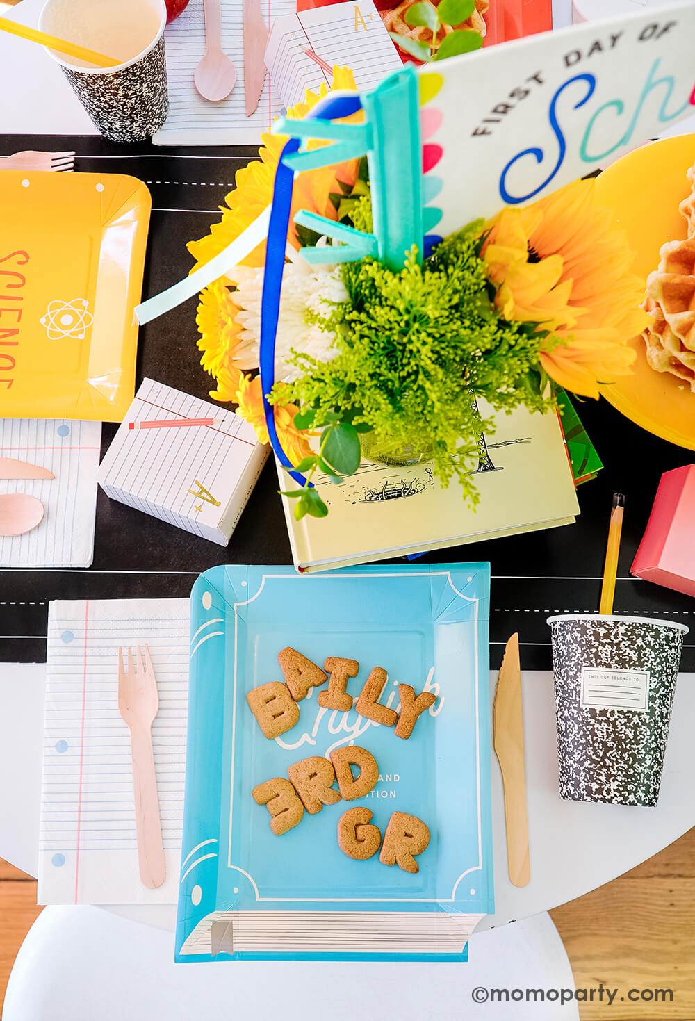 An overhead shot of a first day of school celebration table featuring multiple school themed party goods from Momo Party, including text book shaped plates in different colors, eraser shaped treat boxes, notebook napkins, composition party cups with pencil straws, all on the chalkboard table runner. With stacked waffles, treats, apples, alphabet cookies spelling Baily 3rd Grade on the textbook shaped plate, this makes a perfect idea for your child's back to school party or first day of school celebration!
