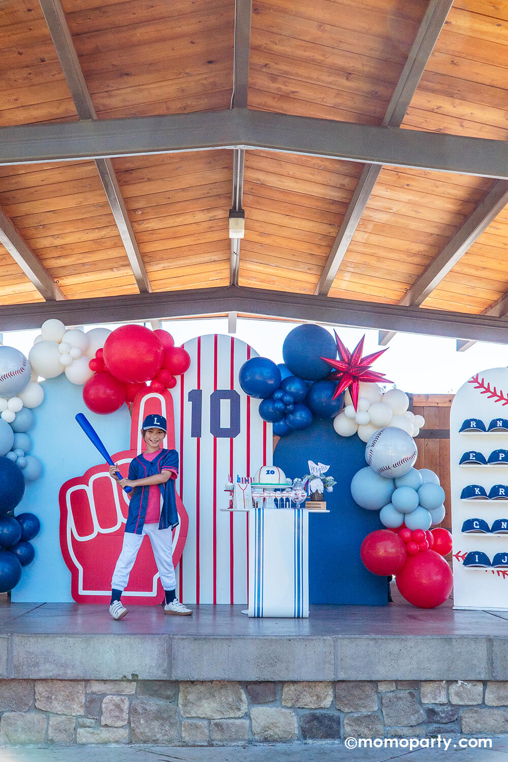 Child in a baseball-themed costume standing in front of a colorful baseball themed party backdrop with festive balloons and decorations styled by Momo Party.
