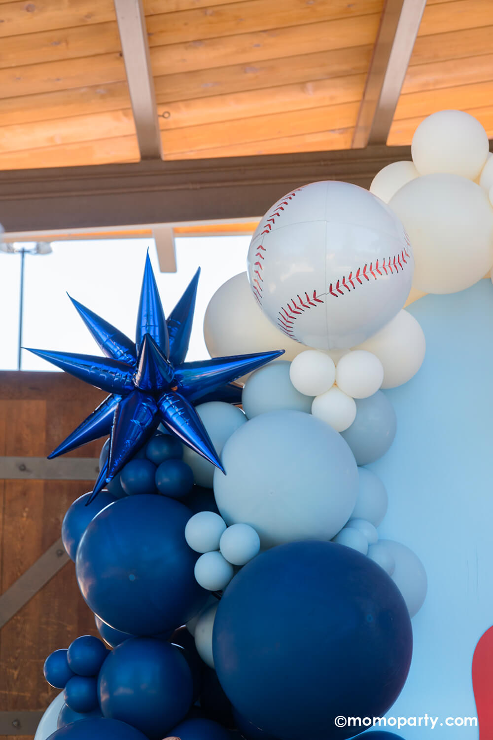 A kid's baseball themed birthday party backdrop decorated with a balloon garland in the classic baseball colors of blue, red and white adorned with a blue starburst foil balloon and a baseball shaped orbz foil balloon from Momo Party.
