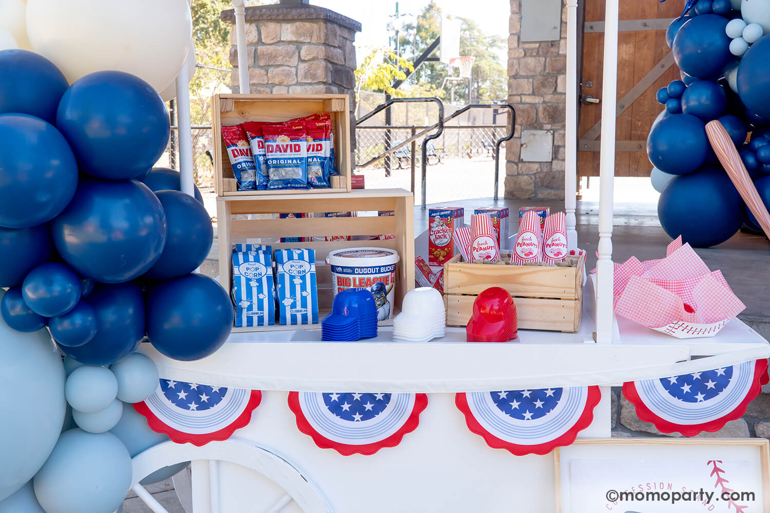 A candy cart decorated with baseball themed balloons in red, blue and white and all-star bunting garland by Momo Party filled with classic baseball ballpark snacks includingCracker Jacks, bags of sunflower seeds, Big League Chew gum balls in a bucket, popcorn in blue striped bags in wooden crates and a box of peanuts, along with mini baseball helmet shaped cups for ice cream and treats, a nostalgic yet with a modern twist set up for baseball snack display at a kid's baseball themed birthday party.