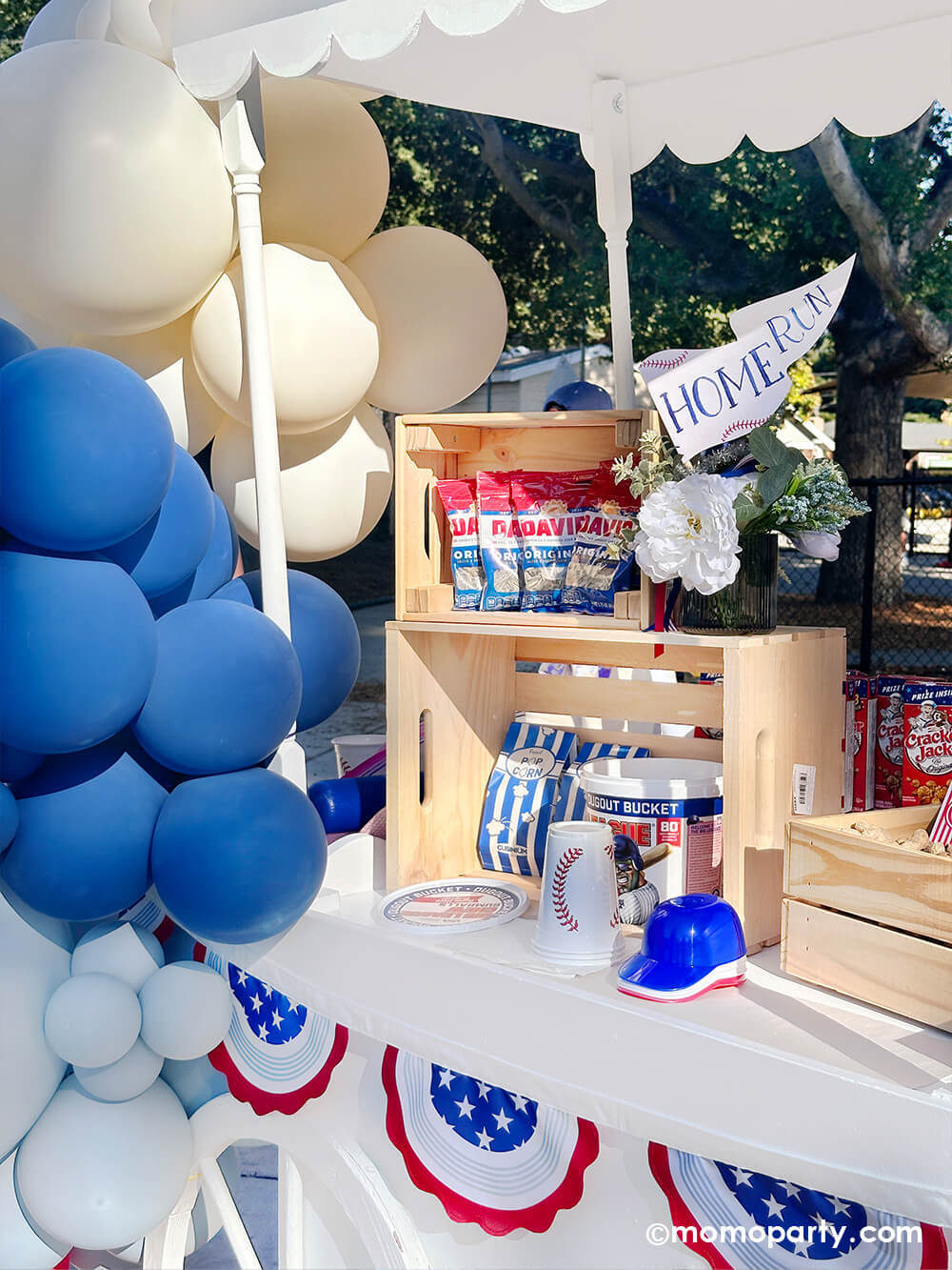 A candy cart decorated with balloon garland and bunting garland, filled with classic baseball ballpark snacks including sunflower seeds, popcorn, Cracker Jacks, and Big League gum balls in wooden crates and a vase with Home Run party pennant from Momo Party at a kid's baseball themed party.