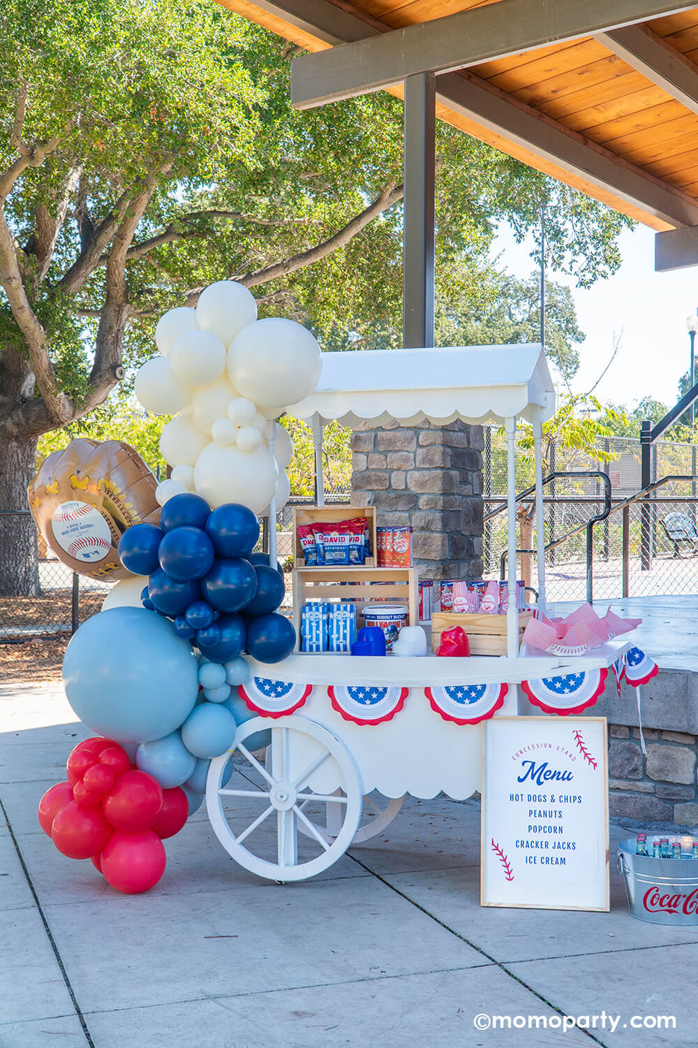A candy cart decorated with baseball themed balloons in red, blue and white and all-star bunting garland by Momo Party filled with classic baseball ballpark snacks includingCracker Jacks, bags of sunflower seeds, Big League Chew gum balls in a bucket, popcorn in blue striped bags in wooden crates and a box of peanuts, along with mini baseball helmet shaped cups for ice cream and treats, a nostalgic yet with a modern twist set up for baseball snack display at a kid's baseball themed birthday party.