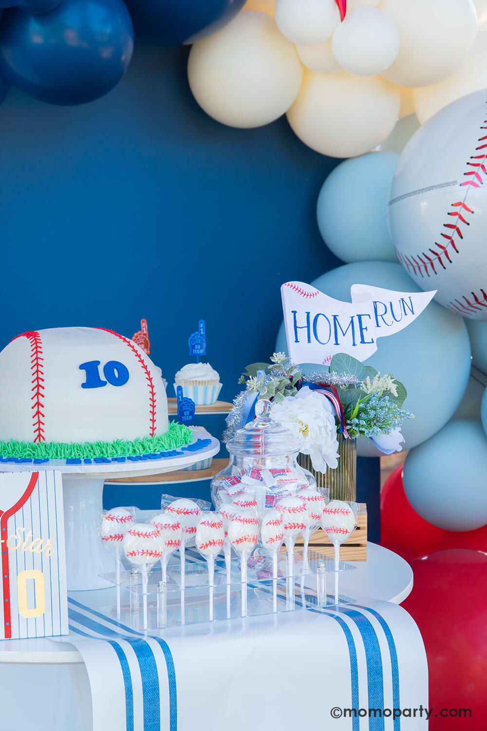 A baseball themed cake and dessert table against a navu backdrop styled by Momo Party featuring a baseball shaped buttercream cake with number 10 on it, cupcakes topped with foam finger toppers, baseball lollipops and a candy jar with Big League Chews gum balls, with a floral bouquet and a "Home Run" party pennant from Momo Party as the centerpiece.