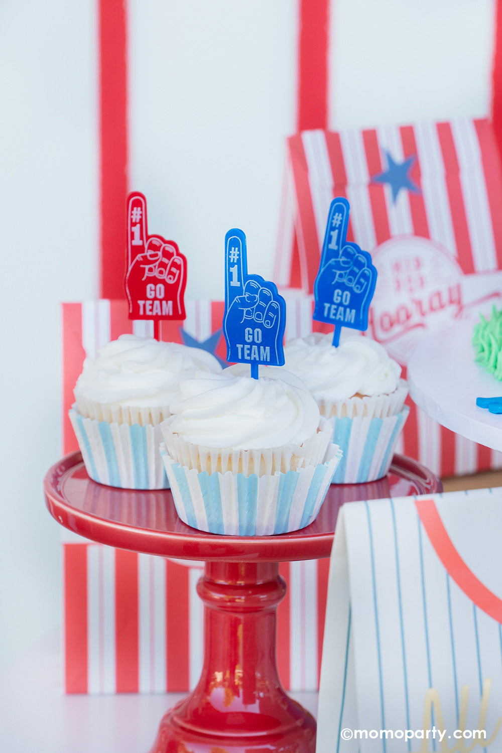 A baseball themed cake and dessert table against a baseball pinstripe backdrop styled by Momo Party featuring the Hip Hip Hooray red striped goodie bags, cupcakes topped with GO TEAM toppers in blue and red on a red cupcake stand and baseball pinstripe treat bag.