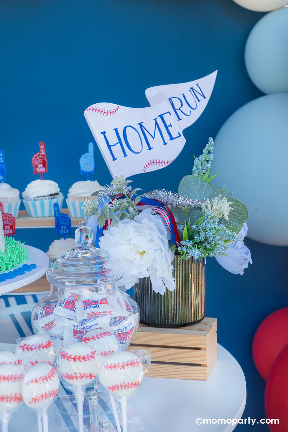 A baseball themed cake and dessert table against a navy backdrop styled by Momo Party featuring a baseball shaped buttercream cake with number 10 on it, cupcakes topped with foam finger toppers, baseball lollipops and a candy jar with Big League Chews gum balls, with a floral bouquet and a "Home Run" party pennant from Momo Party as the centerpiece.