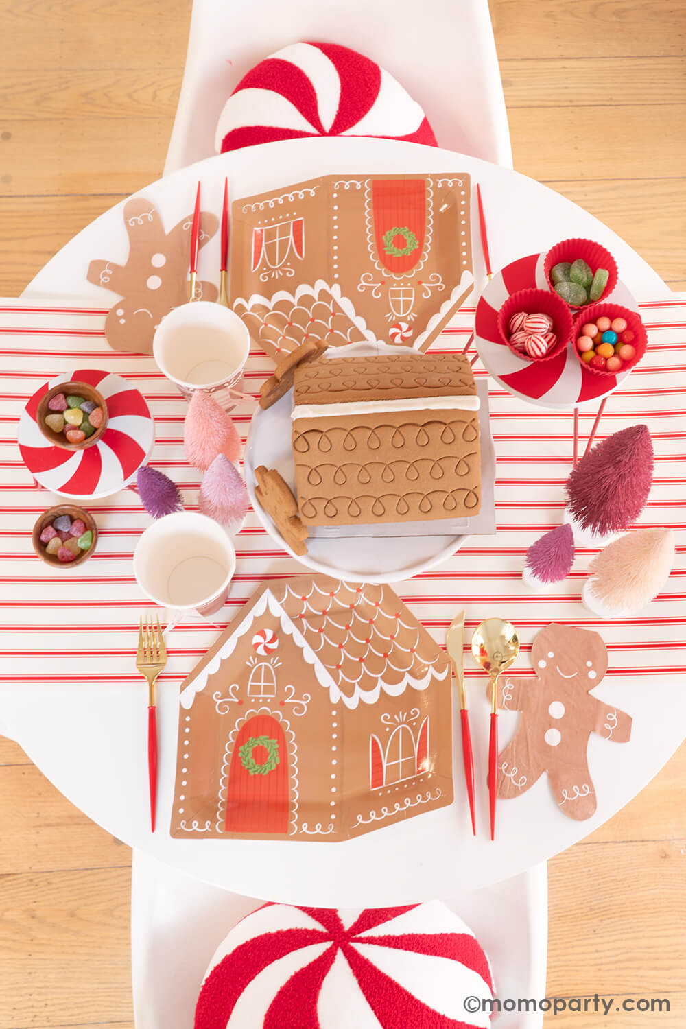 Top view of Momo Party's Gingerbread House Decoration Home Party: A dining table adorned with My Mind's Eye Gingerbread House-Shaped Plates, Smiley Gingerbread Man Napkins, Gold and Red Cutlery Sets, and Gingerbread House Paper Party Cups with Handles. A pre-built Gingerbread House sits in the center, ready for decorating, surrounded by Christmas Bottle Brush Trees and candies—all atop the festive Believe Christmas Red Striped Table Runner. What a delightful way to celebrate the holiday with kids at home!