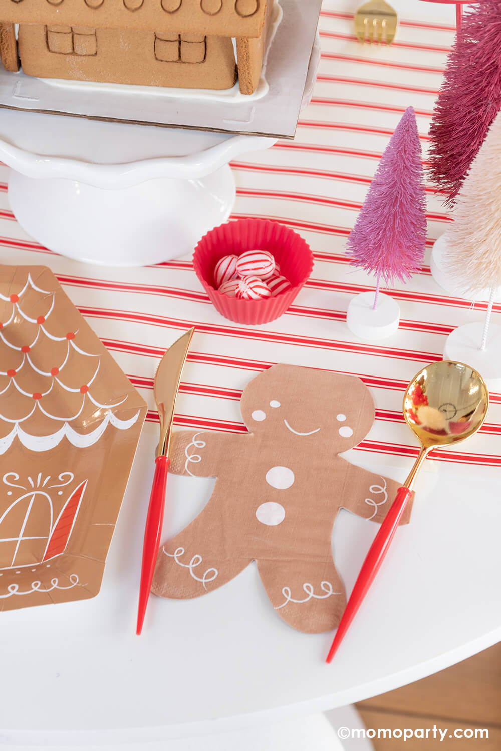 A corner of a festive holiday party table with Momo Party's smiley gingerbread man shaped napkin and festive red and gold cutlery placed on the sides on his hands next to a gingerbread house shaped plate which makes a playful tablescape. Around it is some pink bottle wash Christmas tress in different shades and some peppermint candies ready for decorating the gingerbread house for a kid's holiday fun activity. 