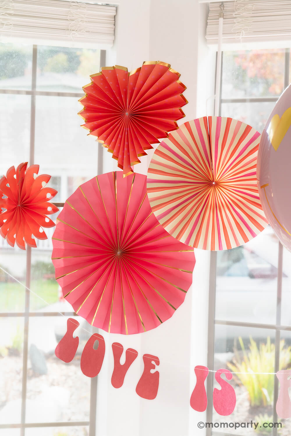 A close-up of Momo Party's Groovy Valentine's Day Party decorations, showcasing the My Mind's Eye Heart You Paper Fan Set with both round and heart-shaped paper fans. The set includes 6 assorted paper fans, adding a delightful touch to the celebration. Paired with a retro-style Love Is All You Need Banner hanging below, these products offer an easy and quick setup for the perfect Valentine's Day party or any romantic occasion. A perfect inspiration for your V-day celebration!"