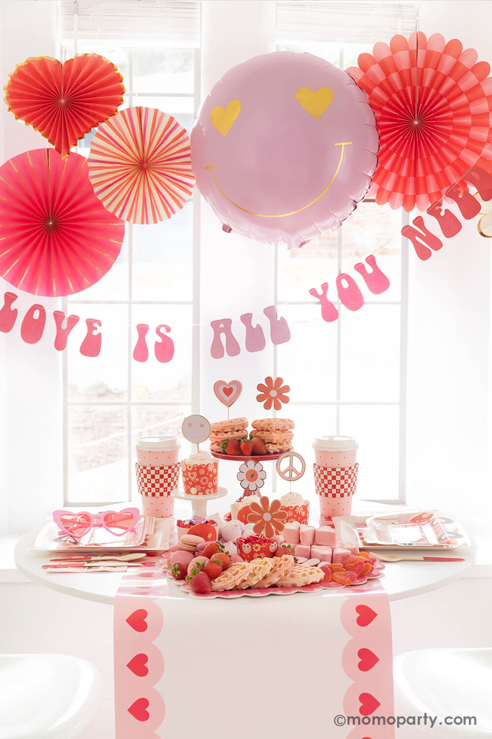 A Valentine's Day celebration featuring Momo Party's Groovy Valentine's Day Party box set up with pink and red paper fans in heart shape and a "love is all you need" banner in retro type hung below the paper fans. On the table you can see a sweet board featuring treats and snacks in red and pink colors including heart shaped waffles, strawberries, macarons, marshmallows etc. Next to the desert board featured retro inspired Valentine's plates, party cups and napkins, a perfect inspo for Vday celebration!