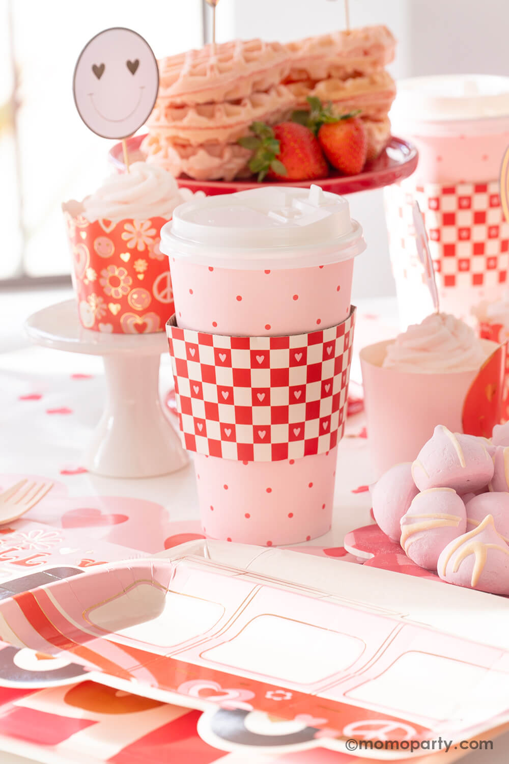 A sweet close-up of Momo Party's Groovy Valentine's Party table, featuring My Mind's Eye's Valentine's party supplies in a retro groovy style. There is Red Checks To-Go Cup wrapped in lively red checks with a mini heart pattern, alongside Occasions by Shakira - Luv Bus Paper Plates. Cupcakes with baking cup and topper, heart-shaped wafers on a red cake stand add a delightful touch, all on the Pink Heart Border Paper Table Runner, create a perfect inspiration for a fun Valentine's celebration