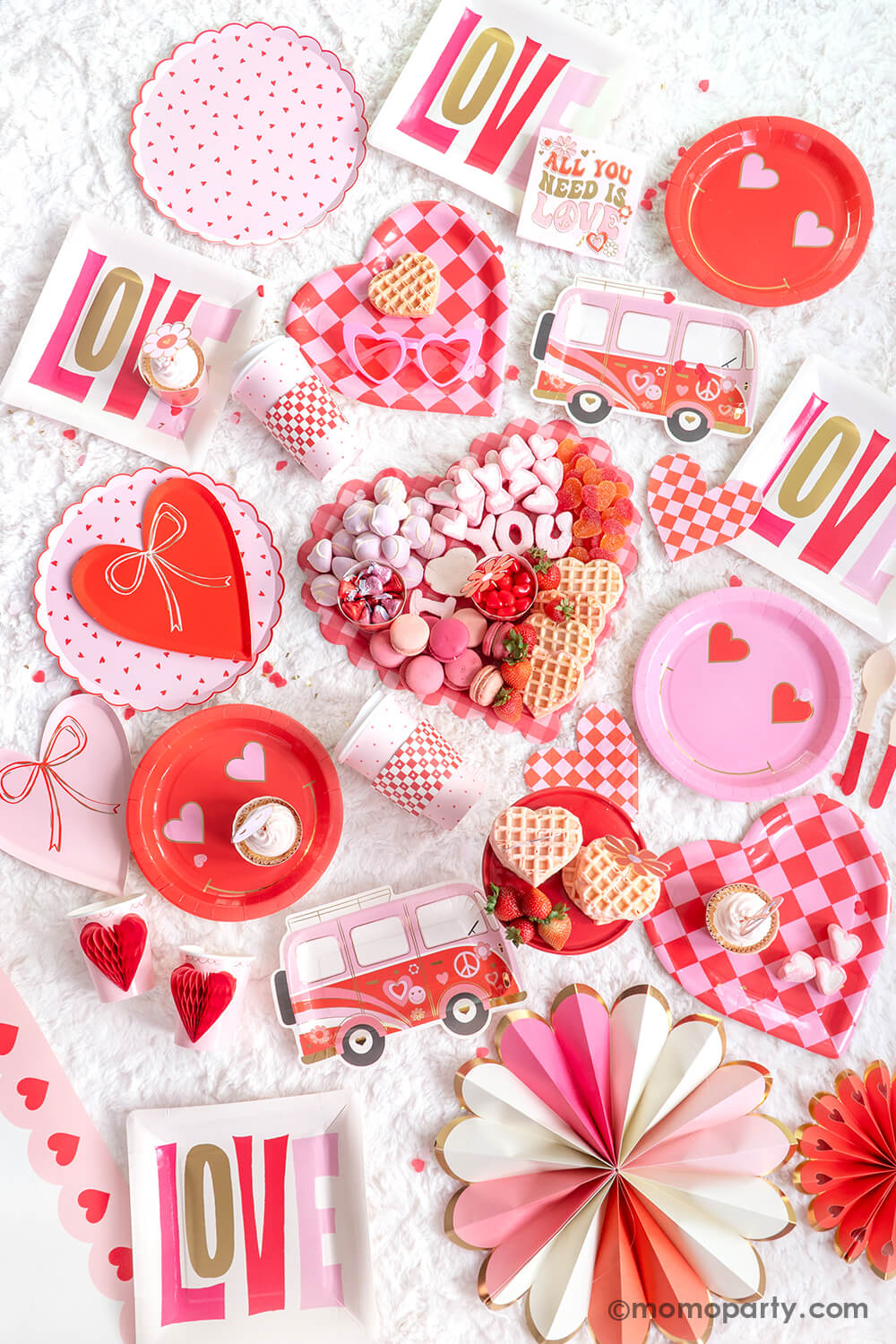Top view displayed Momo Party's 2024 Valentine's Day party collection. Featuring  Heart Pattern Dinner Plates, Heart With Bow Plates, Honeycomb Heart Cups, Occasions by Shakira - Luv Bus Paper Plates, and All You Need is Love Napkins. Also, Checkered Heart-Shaped Paper Plates, Square Love Paper Plates, Heart Eyes Paper Plates, Red Checks To-Go Cup Set, and Pink Heart Border Paper Table Runner. Not to forget the Checkered Heart-Shaped Tray adorned with sweet treats, for a festive Valentine's celebration