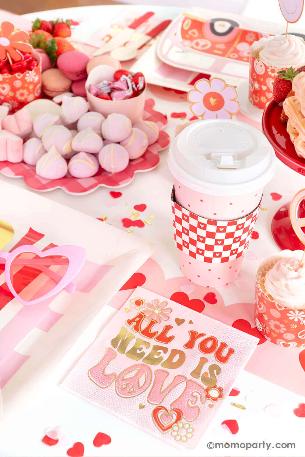 A party table featuring Momo Party's Groovy Valentine's party box with My Mind's Eye's retro Valentine's party supplies including baking cups, "all you need is love" napkins, VW bus shaped plates and red checkered party cups. On the table there's a heart shaped sweet board filled with Valentine's Day themed treats including macarons, strawberries, heart shaped marshmallows, heart shaped pink waffles and candies, a perfect inspo for a fun Valentine's celebration!