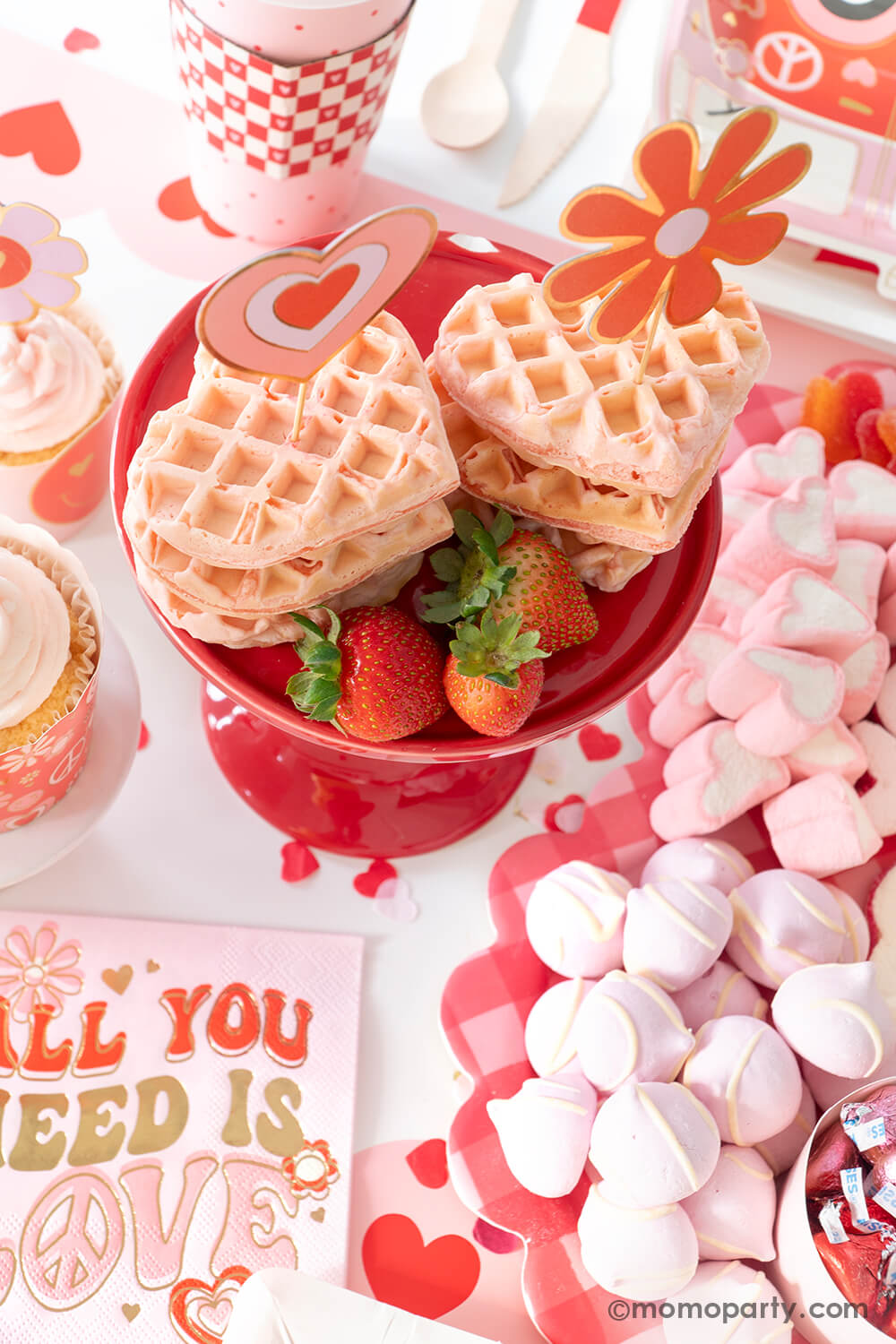 A festive Valentine's Day party table featuring a sweet board filled with heart shaped treats and desserts in pink and red. In the middle some heart shaped pink waffles topped with Momo Party's groovy love themed toppers with strawberries next to them. On the table there are lots of retro inspired Valentine's Day themed party supplies including Momo Party's all you need is love napkins, vw bus shaped plates, and red checkered party cups, makes this a perfect info for a sweet Valentine's Day celebration.