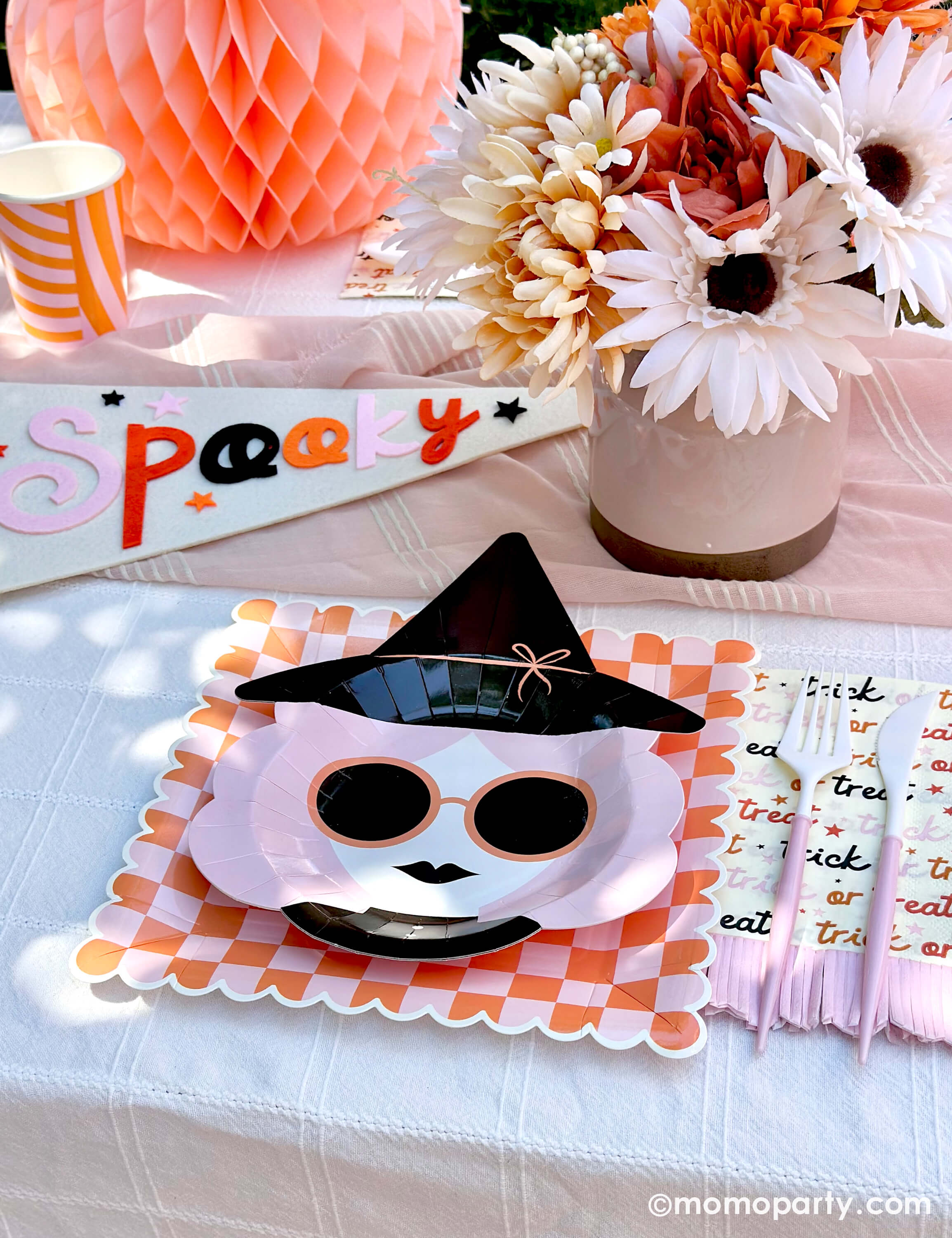 A pink Halloween table featuring Momo Party's pink witch plates paired with orange/pink checkered plates, with boho vibe fall flower arrangement and pastel peach honeycomb pumpkins as the centerpiece, and a felt party pennant that has "spooky" written on, this tablescape gives a great inspiration for a modern and chic Halloween party table decoration ideas.