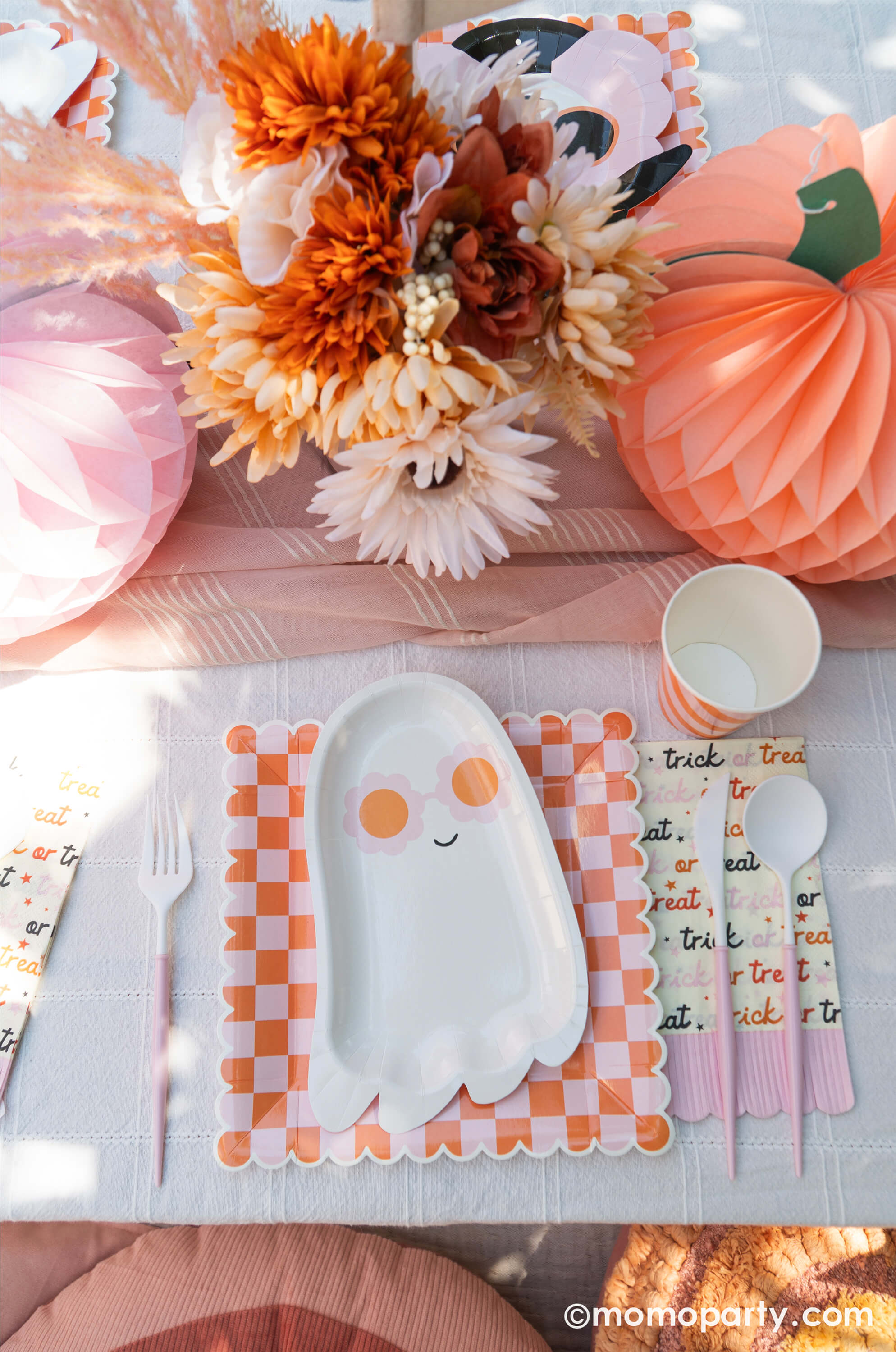 A pink Halloween table featuring Momo Party's pink sunny ghost shaped plates paired with orange/pink checkered plates, with boho vibe fall flower arrangement and pastel peach and pink honeycomb pumpkins as the centerpiece, this tablescape gives a great inspiration for a modern and chic Halloween party table decoration ideas.