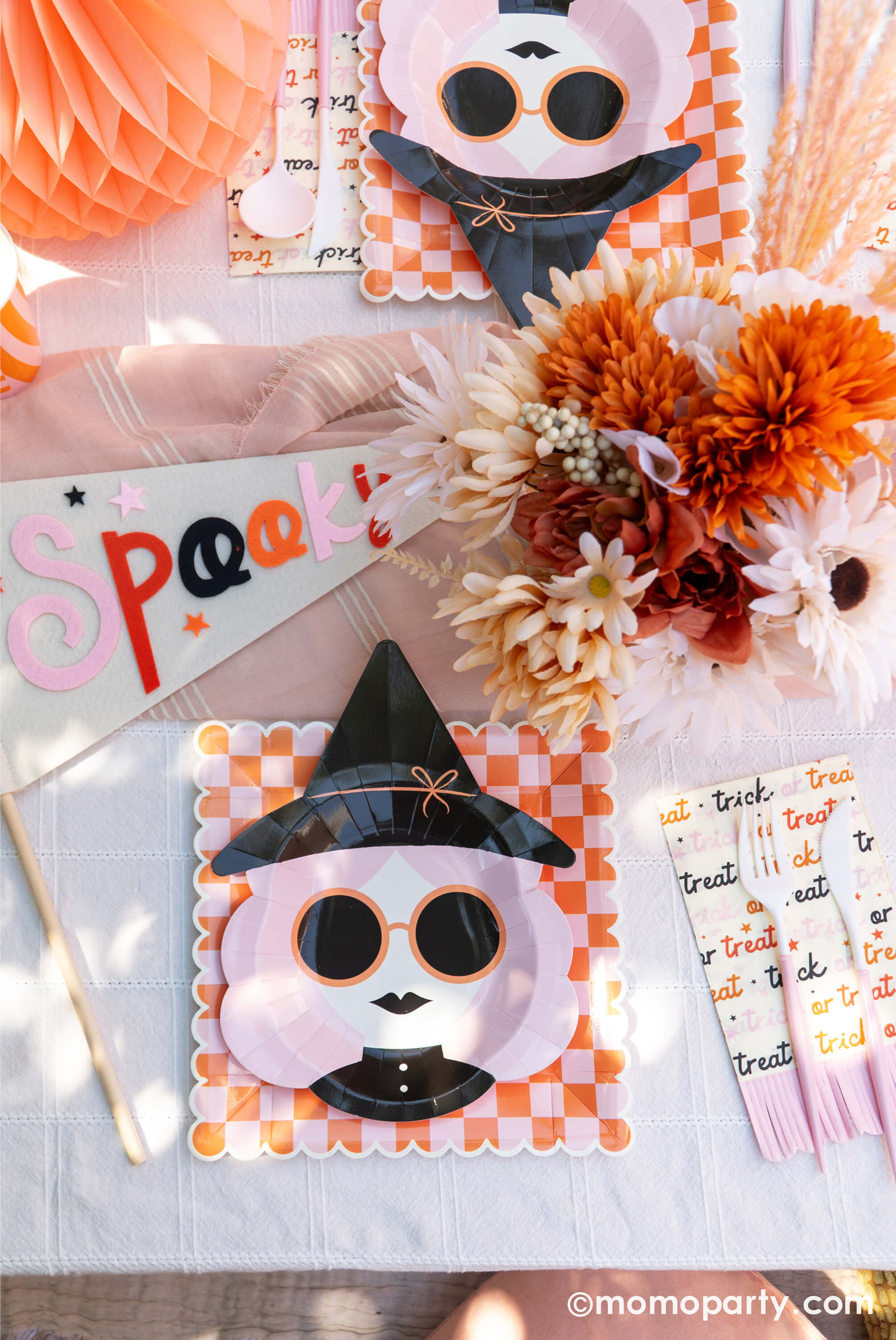 A pink Halloween table featuring Momo Party's pink witch plates paired with orange/pink checkered plates, with boho vibe fall flower arrangement and pastel peach honeycomb pumpkins as the centerpiece, and a felt party pennant that has "spooky" written on, this tablescape gives a great inspiration for a modern and chic Halloween party table decoration ideas.
