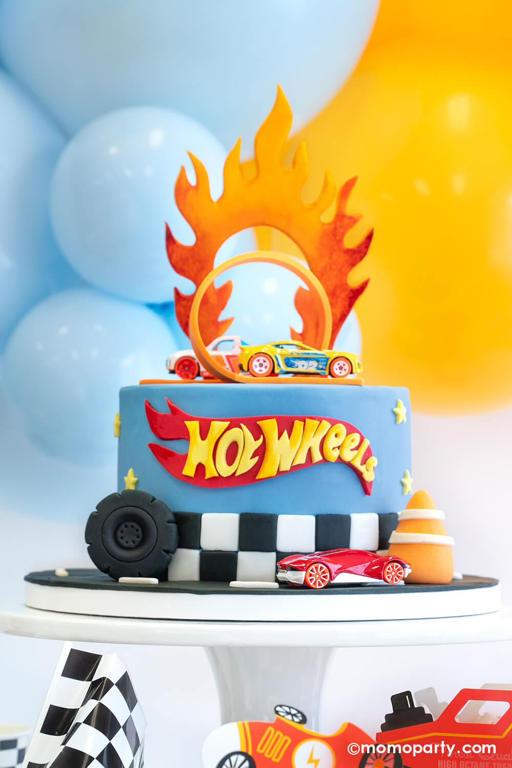 Hot Wheels themed birthday cake with the classic orange curve track and Hot Wheels toy cars by Momo Party. On the side of the cake are a fondant wheel and construction cone, making this a perfect look for kid's Hot Wheels themed birthday party.