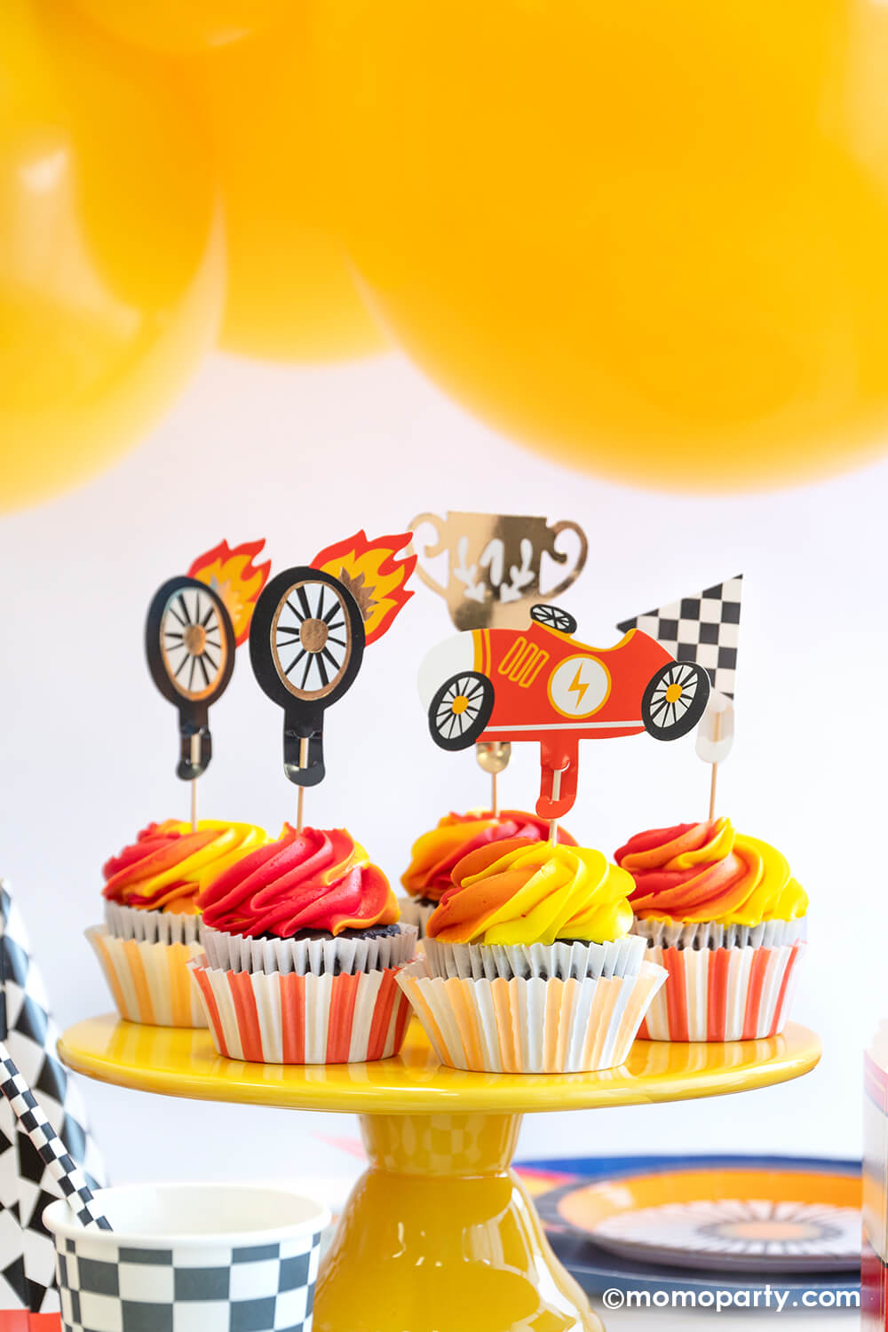 A race car / Hot Wheels themed party featuring orange and yellow swirl cupcakes topped with race car themed toppers including wheels, a vintage race car, a trophy and a checkered flag. In the back there are some yellow party balloons. On the table there are some race car themed tableware including checkered party cups, wheel shaped plates, midnight dinner plates.