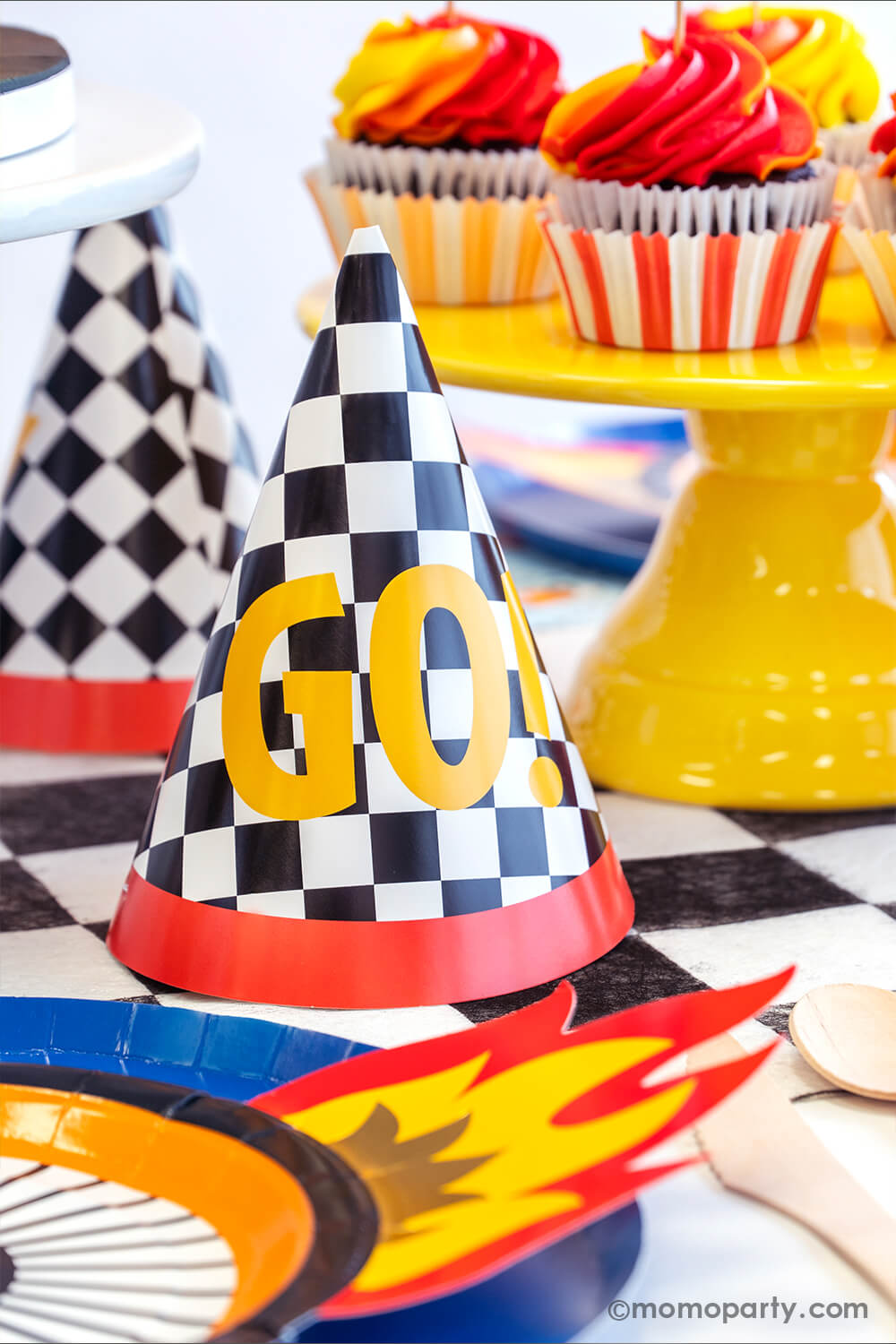Momo Party checkred GO! birthday hats next to red and yellow swirl cupcakes in a Hot Wheels themed birthday party where there's wheel shaped plates with flame on them along with midnight blue dinner plates.