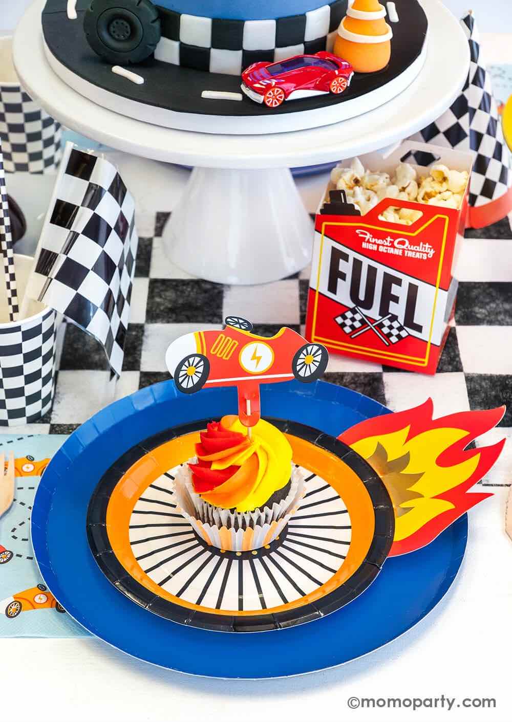 A close up shot of a Hot Wheels themed birthday party table featuring Momo Party's midnight dinner plates with wheel shaped plates, checkered table runner, party cups, paper straws and party cups, Fuel gas shaped treat box with popcorn and blue race car napkins. On the plates there's a orange and red swirl cupcake topped with vintage race car topper - a perfect inspo for boy's Hot Wheels themed birthday party.