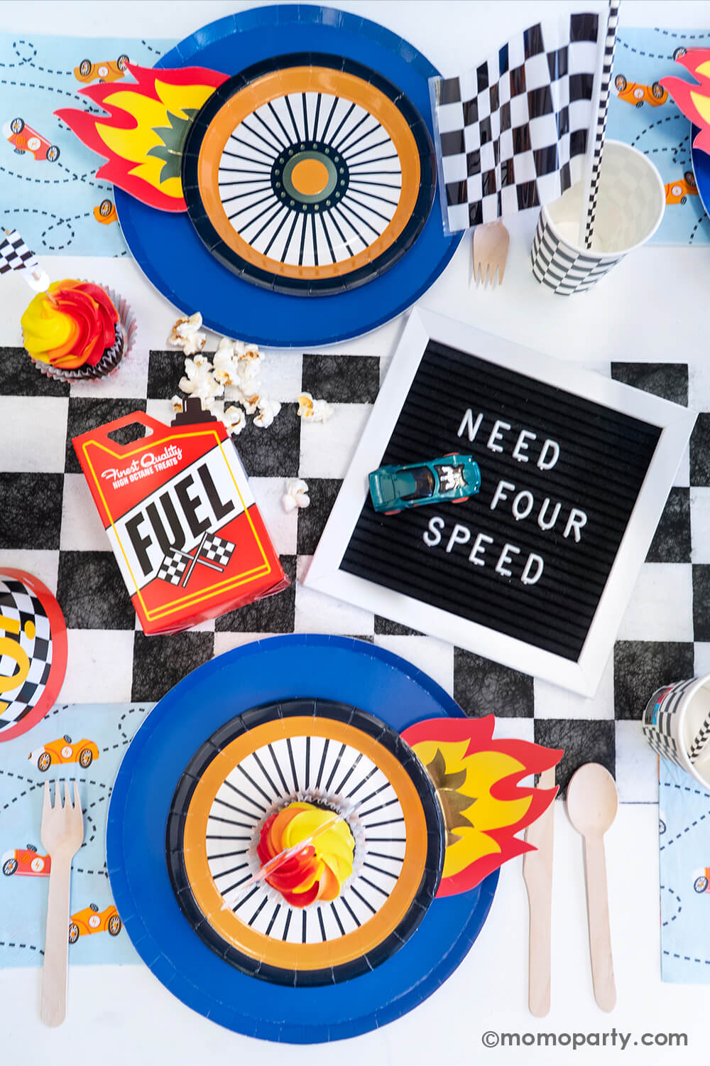 A flatlay shot of a festive "Need FOUR Speed" Hot Wheels themed fourth birthday party table featuring Momo Party's midnight dinner plates with wheel shaped plates, checkered table runner, party cups, paper straws, birthday hats, Fuel gas shaped treat box with popcorn and blue race car napkins . In the middle of the table, there's a black letterboard spelling "NEED FOR SPEED" and a Hot Wheels toy car next to it - a perfect inspo for boy's 4th birthday party.