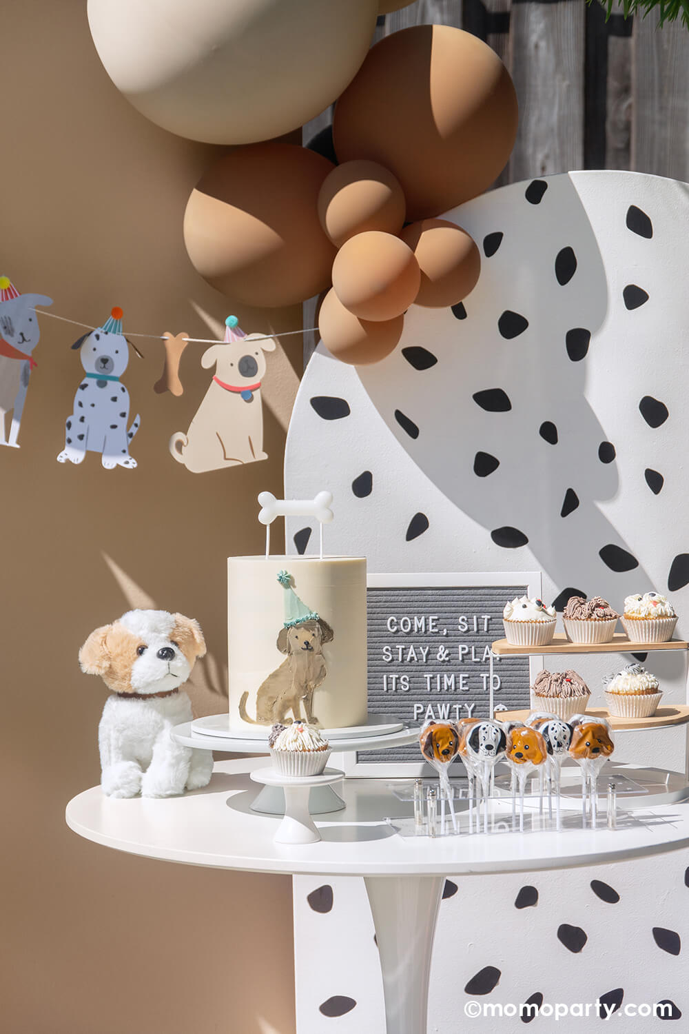 A puppy dog themed kid's birthday party by Momo Party featuring a Dalmatian print backdrop adorned with stunning balloon garland in brown, neutral colors and Meri Meri's puppy party garland featuring puppy pennants with folded ears. In the front of the backdrop, there's a dessert table filled with dog-inspired treats including a buttercream cake with an adorable dog illustration on the side topped with a bone-shaped candle, dog cupcakes, and dog head shaped lollipops.
