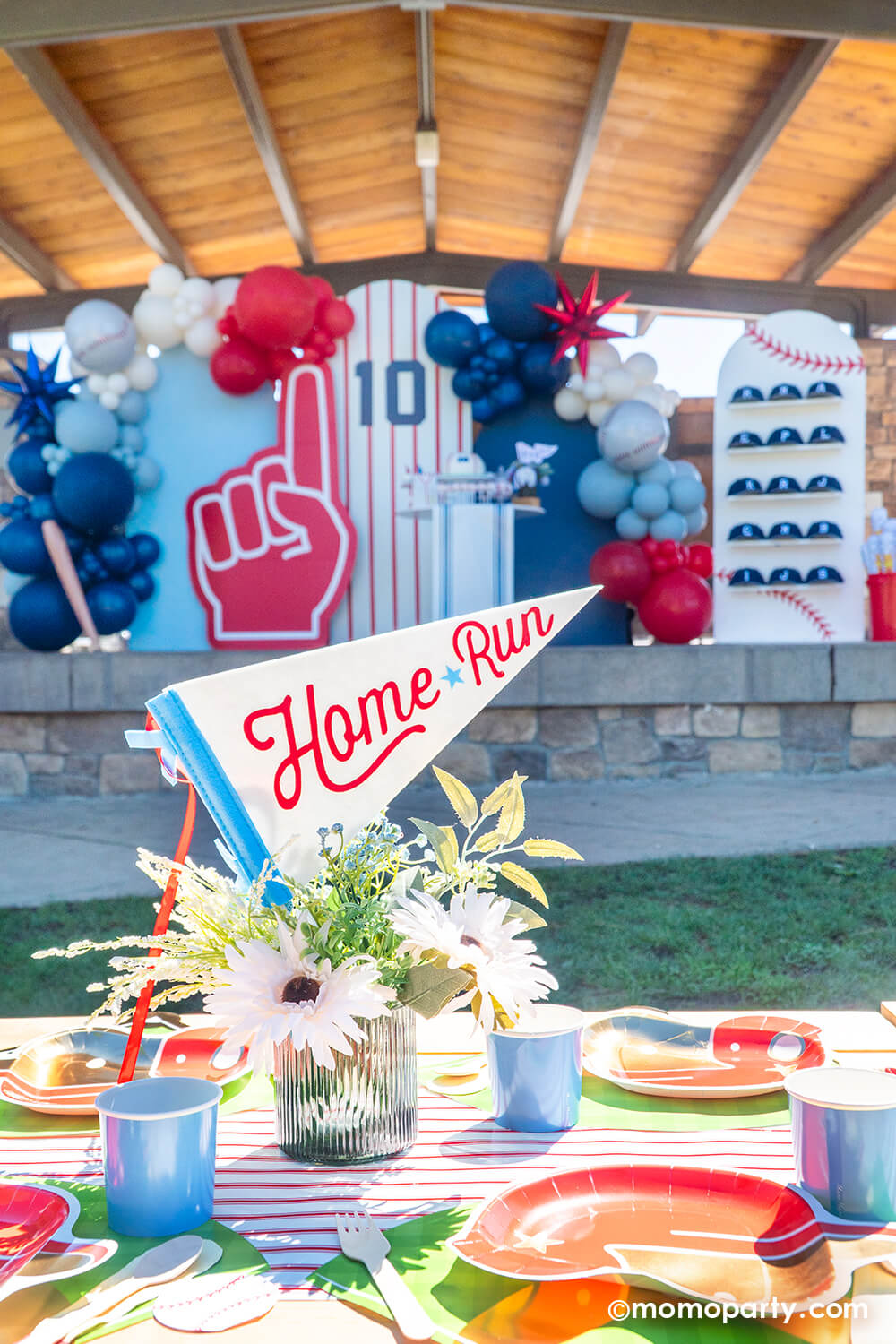 A kid's baseball themed birthday party picnic table setting styled by Momo Party featuring various baseball themed tablewares from Momo Party including baseball helmet shaped plates, cornflower tumbler cups, baseball shaped napkins on the baseball diamond placemats with the red pinstriped table runner underneath. In the center there's a beautiful flower bouquet with the "Home Run" felt party pennant. In the back there's a baseball party backdrop adorned with baseball themed balloons and a cap wall.