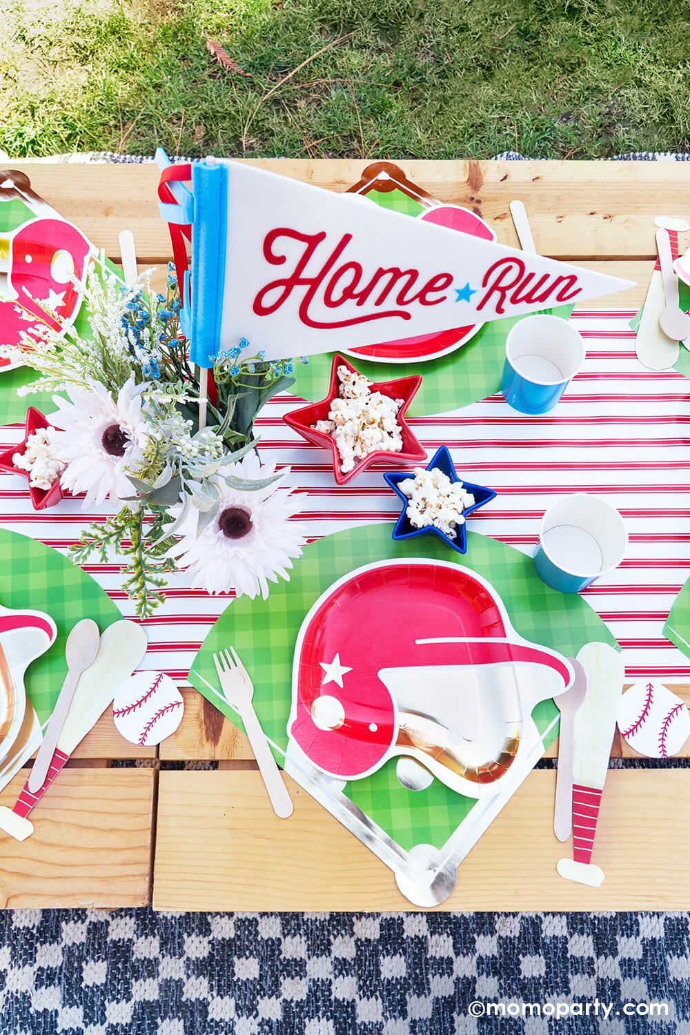 An overhead shot of kid's baseball themed birthday party picnic table setting styled by Momo Party featuring various baseball themed tablewares from Momo Party including baseball helmet shaped plate, cornflower tumbler cups, baseball shaped napkins on the baseball diamond placemats with the red pinstriped runner underneath. In the center there's a beautiful flower bouquet with the "Home Run" felt party pennant and some star shaped trays in blue and red filled with popcorn.