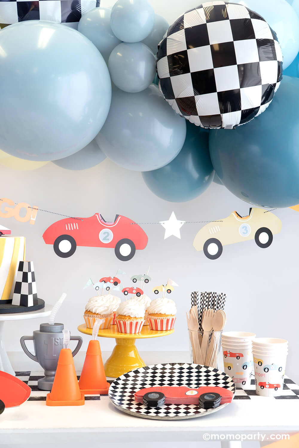A festive race car themed party set up by Momo Party featuring a blue balloon garland adorned with checkered foil balloons, underneath hung a vintage race car party garland. On the table are some cupcakes topped with race car toppers next to a couple cone shaped and trophy shaped sippers. With checkered dinner plates, table runner and party straws, along with red race car napkins and party cups, makes a perfect inspiration for a kid's "Two Fast" second birthday party.