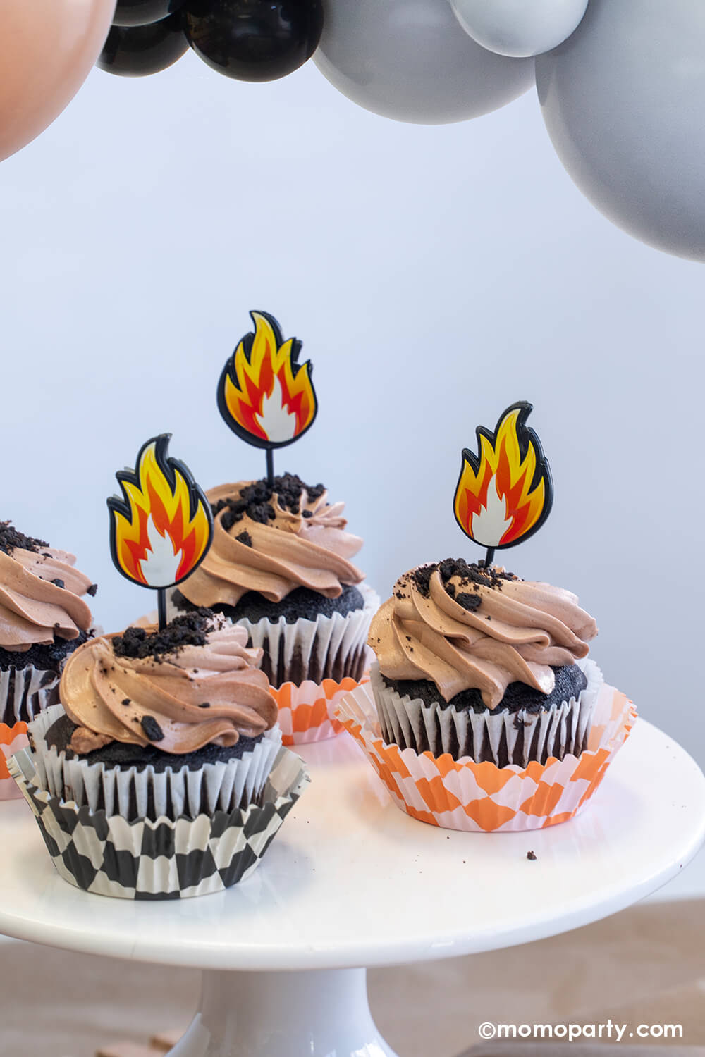 A close-up shot of a monster truck themed party decorated with party balloon garland by Momo Party. The table features a few cupcakes wrapped with orange and black checker patterned wraps with brown icing and oreo crumbs on them, each of them is topped with a fire flame topper. Making these cupcakes a fun treat idea for kid's monster truck themed birthday celebrations!