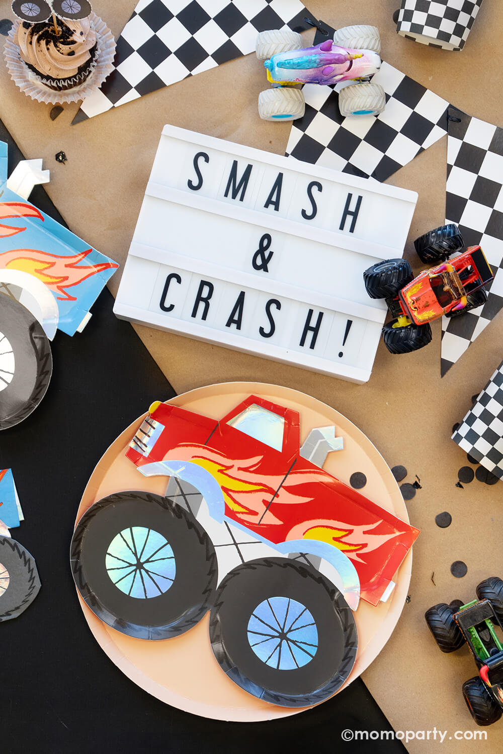 A close up of an overhead shot of Momo Party's Monster Truck Party Collection featuring various monster truck themed tableware and decors including a race car flag banner, black, sand and checkered round dinner plates, party cups, straws and monster truck shaped plates and napkins, with monster truck shaped cupcake kit and "Smash & Crash" sign, monster truck toys on a brown craft paper runner, this collection is perfect for your little monster truck fan's birthday celebration!