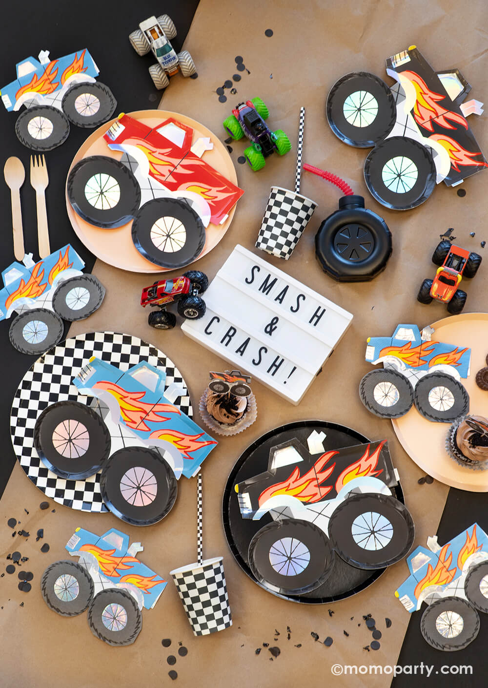 A overhead shot of a monster truck themed party tabescape featuring various monster truck themed tableware from Momo Party including round black and checkered dinner plates, party cups, straws and monster truck shaped plates and napkins, with a brown cupcake topped with a monster truck topper and "Smash & Crash" sign plus truck toys on a brown craft paper runner, this setup is perfect for your little monster truck fan's birthday celebration!