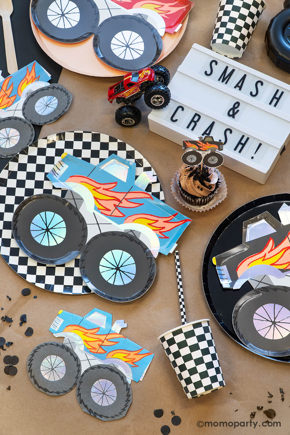 A overhead shot of a monster truck themed party tabescape featuring various monster truck themed tableware from Momo Party including round black and checkered dinner plates, party cups, straws and monster truck shaped plates and napkins, with a brown cupcake topped with a monster truck topper and "Smash & Crash" sign plus truck toys, this setup is perfect for your little monster truck fan's birthday celebration!