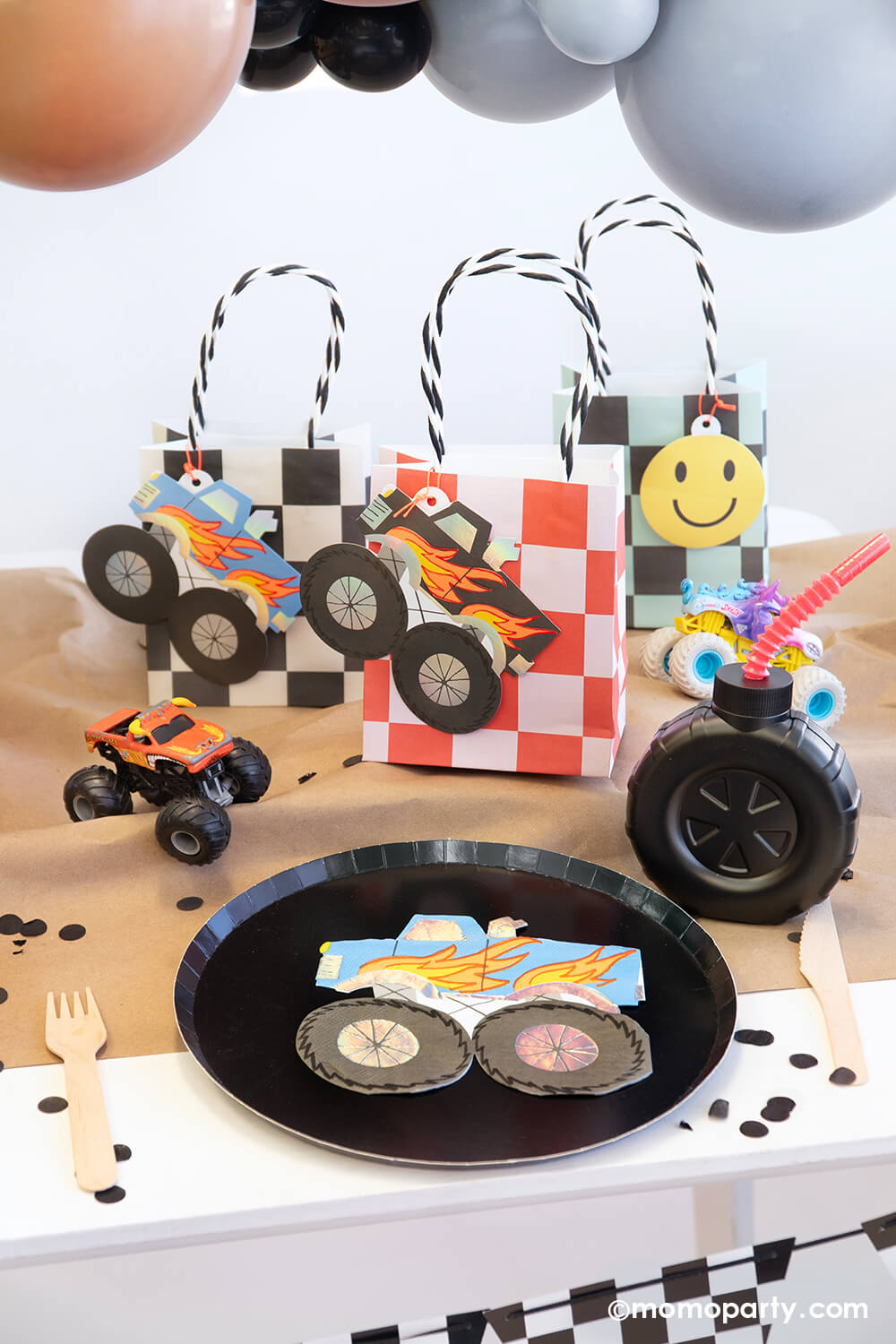 A Monster Truck themed Party Table styled by Momo Party featuring a black round paper dinner plate with a blue monster truck shaped napkin on it, next to them is a big tire shaped tumbler cups with a red plastic straw. Sitting behind are three checker board party bags with monster truck shaped tag and a smiley face tag, all on a brown kraft table runner decorated with monster truck toys and black confetti - a perfect table setting for a fun kid's monster truck party!