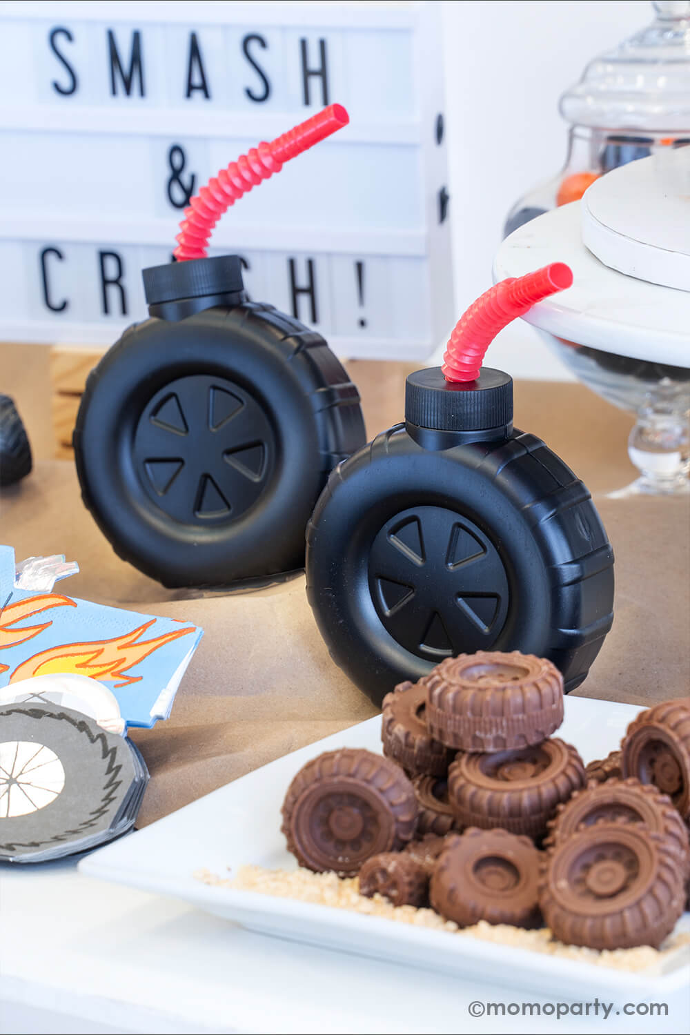 A monster truck themed party table setting styled by Momo Party. In the front there's a plate full off tire shaped chocolate candies and some graham cracker crumbs. The table is covered with brown kraft paper a couple of tire shaped tumbler cups with red straws. In the back there's a sign with "Smash & Crash" - setting up a perfect scene for a fun monster truck themed party for kids.