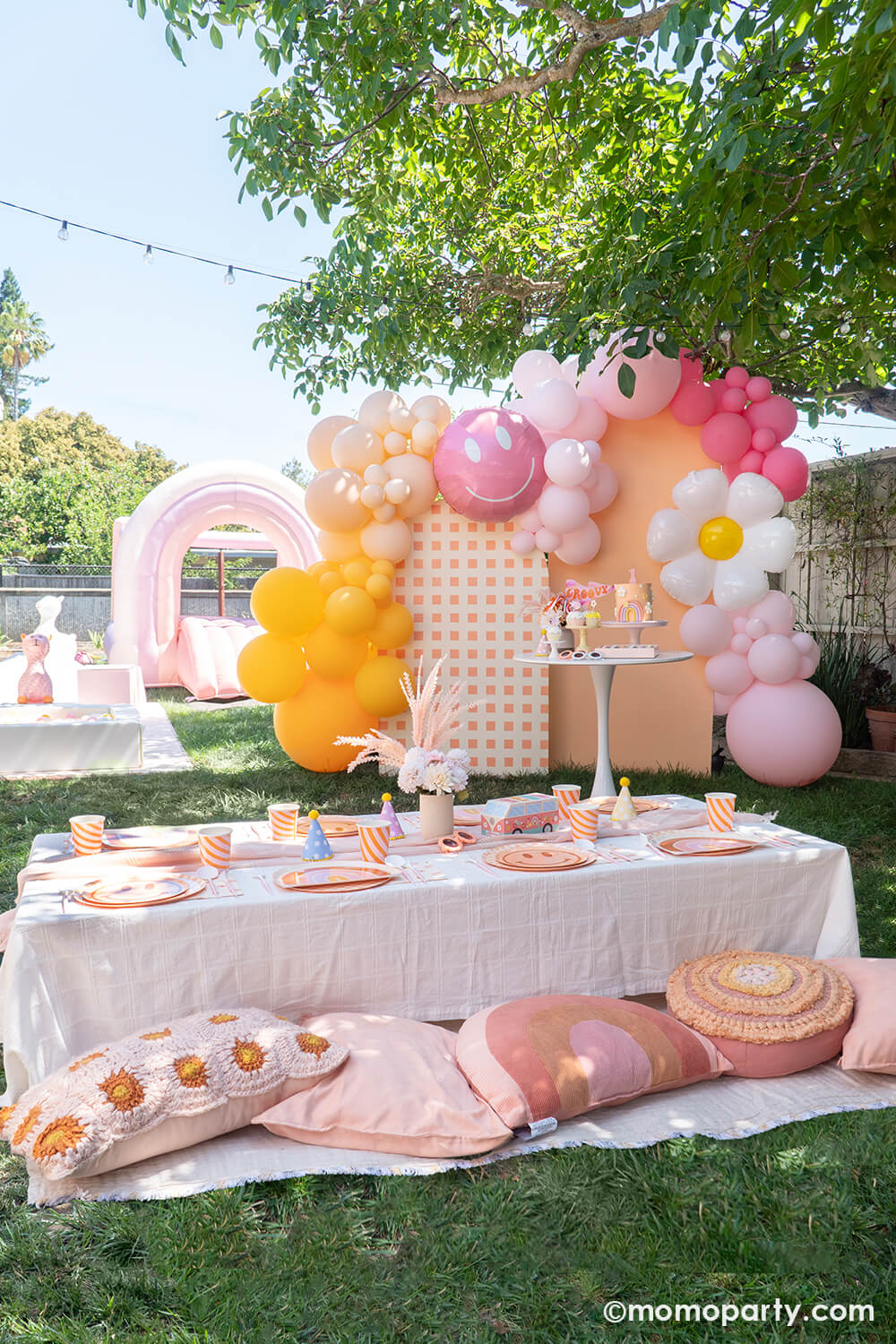 Momo Party One Groovy Baby First Birthday Party, with a birthday girl wearing a Retro Daisy Party Hat, sitting in the hair chair, eating her smash cake with a check patterned wooden Backdrop board, 30" Rosy Smiley face foil balloon by Tuftex Balloons, latex balloon decorations in peach, pink, Goldenrod colors. This decoration is such a stand out backdrop and photo booth for a Groovy birthday parties, baby showers, and most any other joyful event