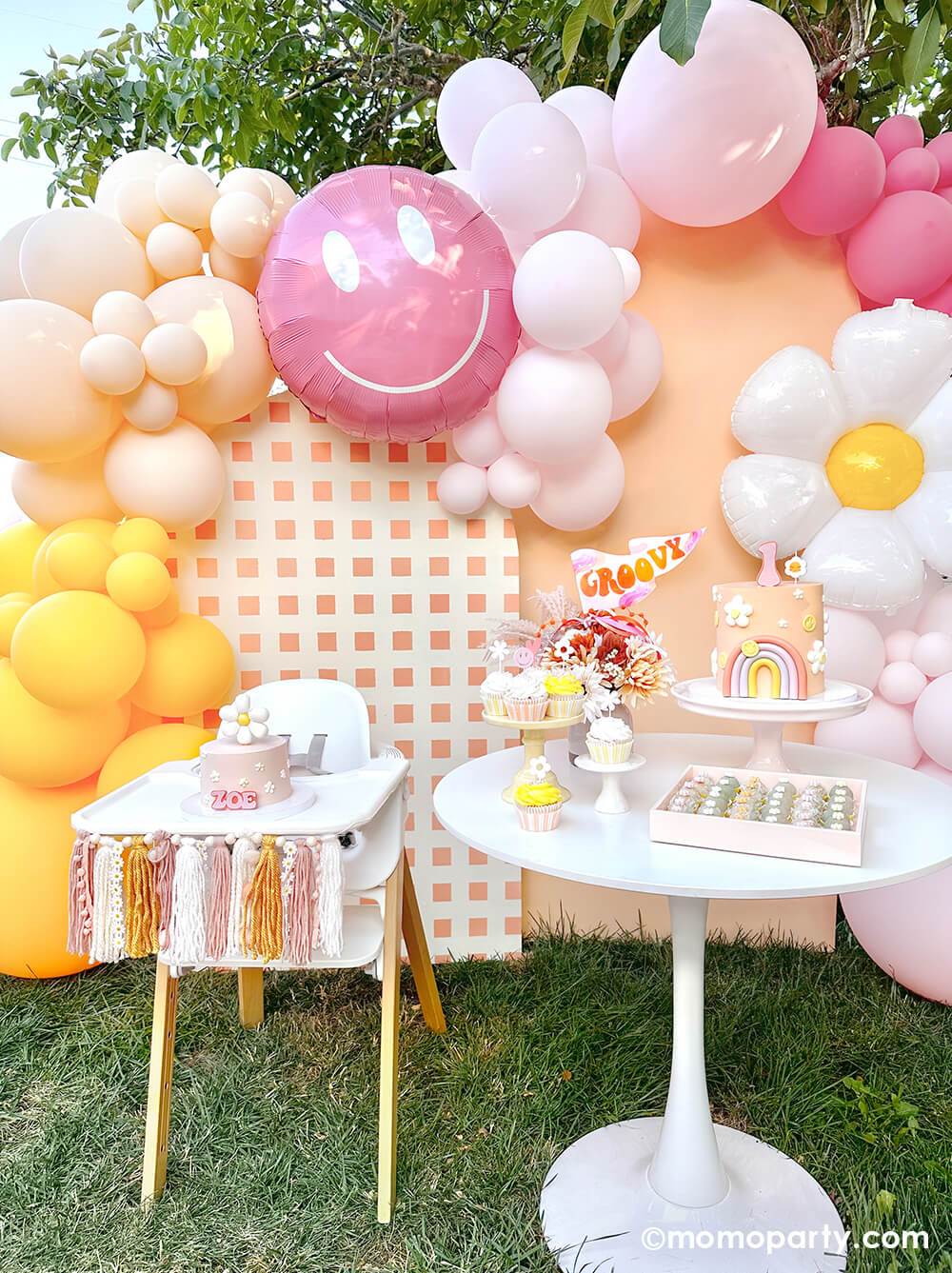 Momo Party’s “One Groovy Baby” First Birthday Party. This outdoor backyard party is decorated with a check-patterned wooden Backdrop board, a 30" Rosy Smiley face foil balloon, a Daisy Foil Mylar Balloon, and latex balloon clusters in peach, pink, and Goldenrod colors. A smash cake with a daisy flower on the Stokke high chair, decorated with Boho Daisy Yarn tassel Banner, on the right side there are birthday cake with a number one topper, Daisy Birthday Candle, Groovy Party Pennant inside the flower vase